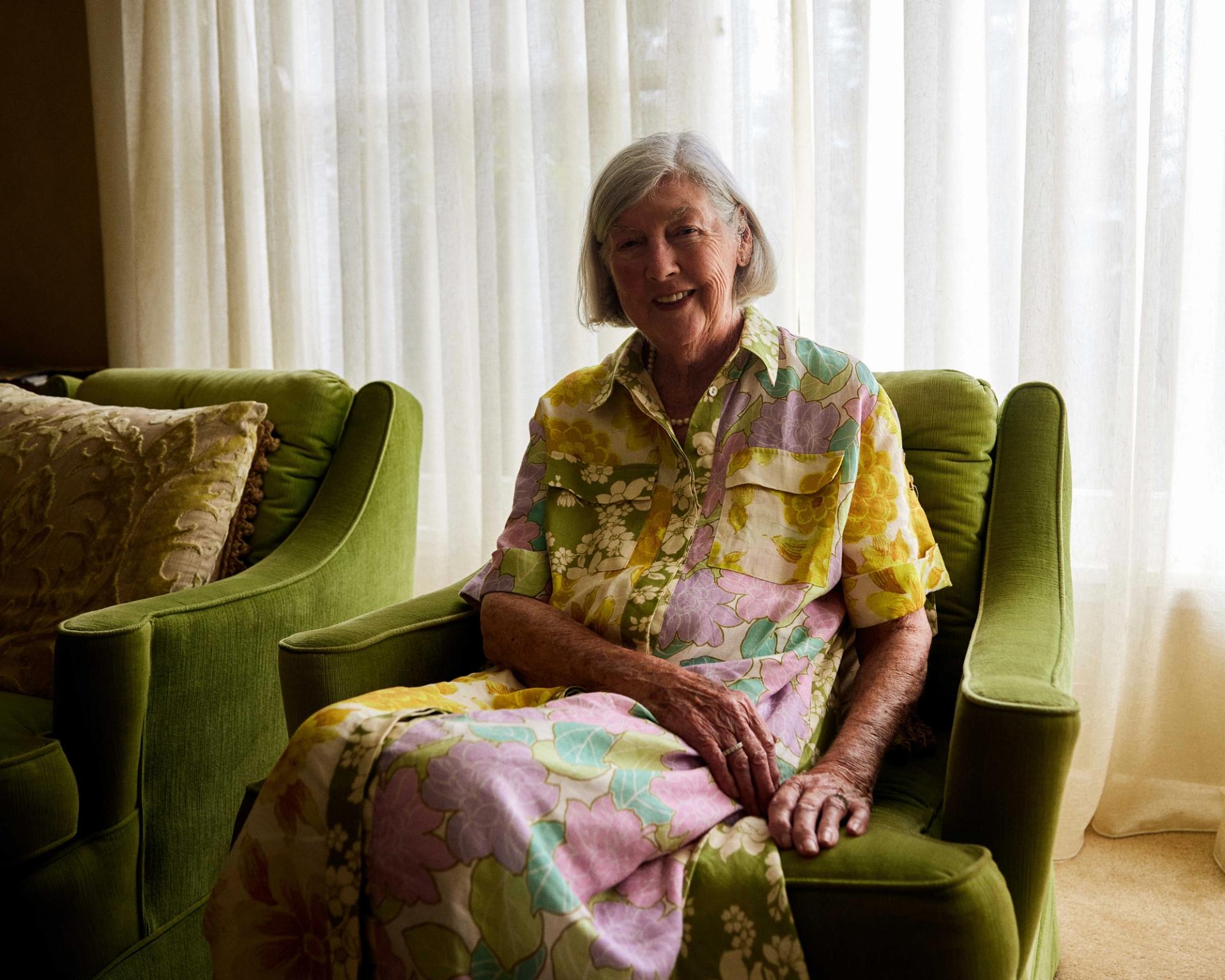 An elderly woman sits on her couch and smiles at the camera.