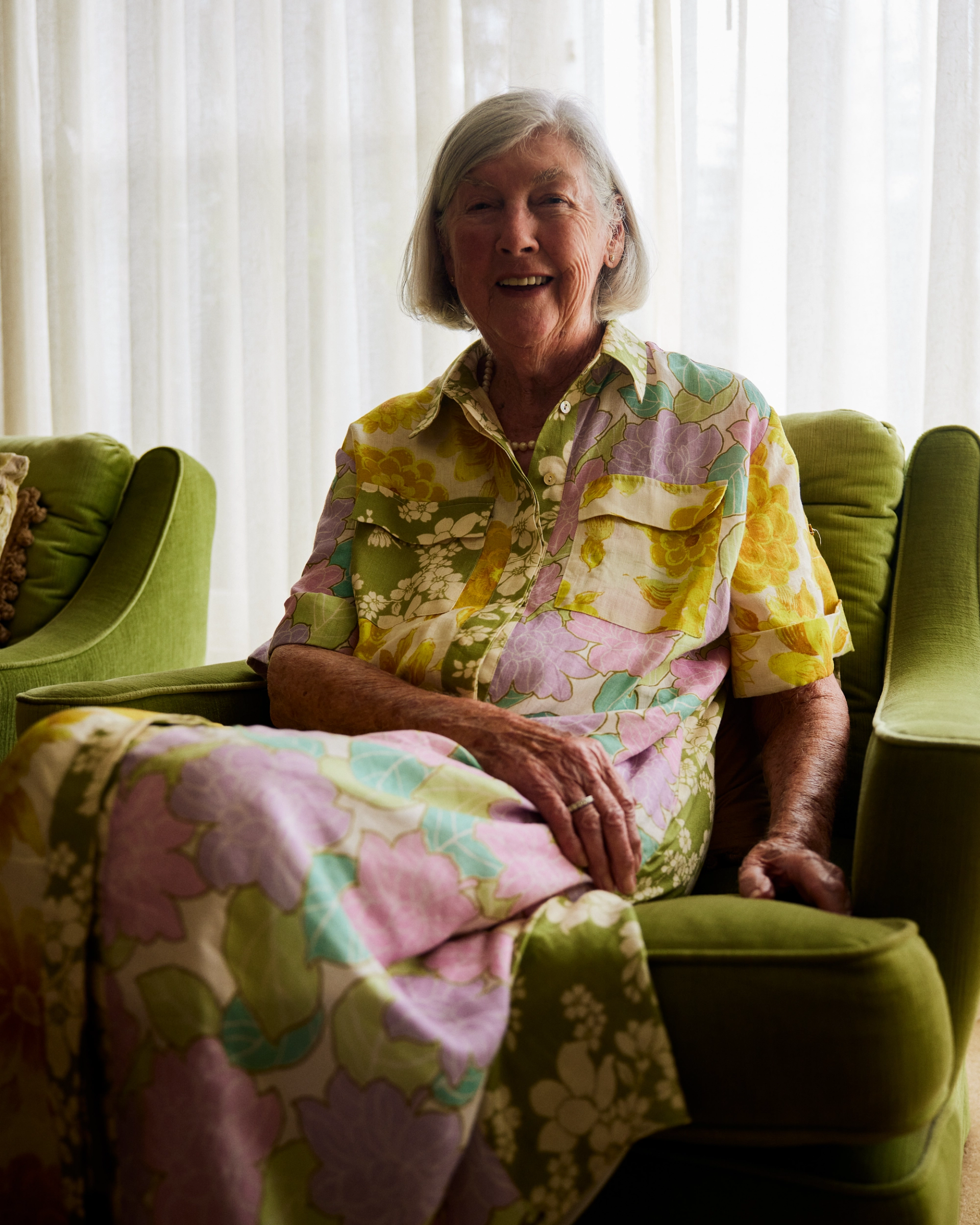 An elderly woman sits on her couch and smiles at the camera.
