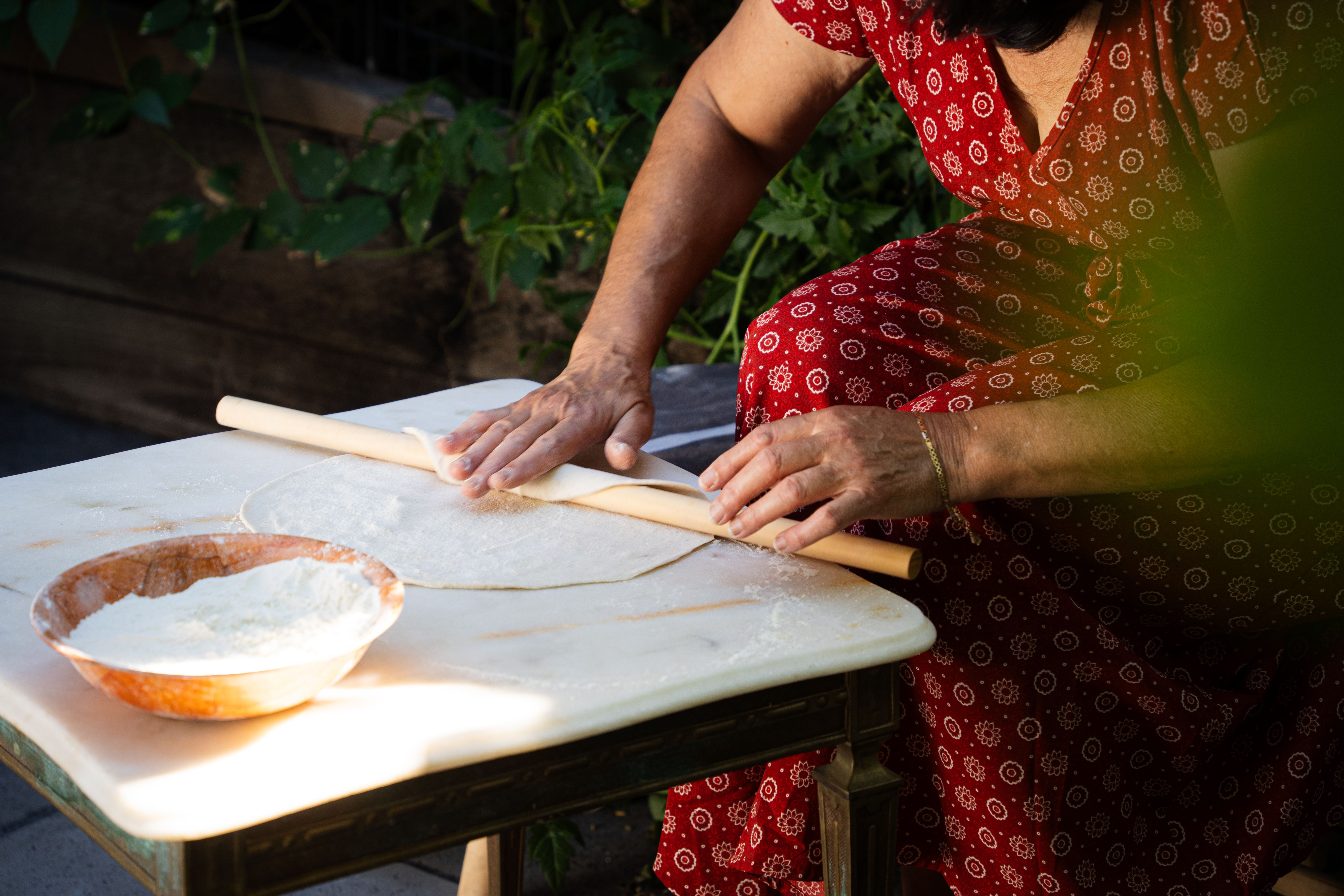 Siham is wearing a red patterned dress and is rolling out dough on a marble slab with a wooden rolling pin. The dough is being shaped into a large, thin circle. She’s rolling it out in her backyard, emphasising a natural and relaxed cooking environment.