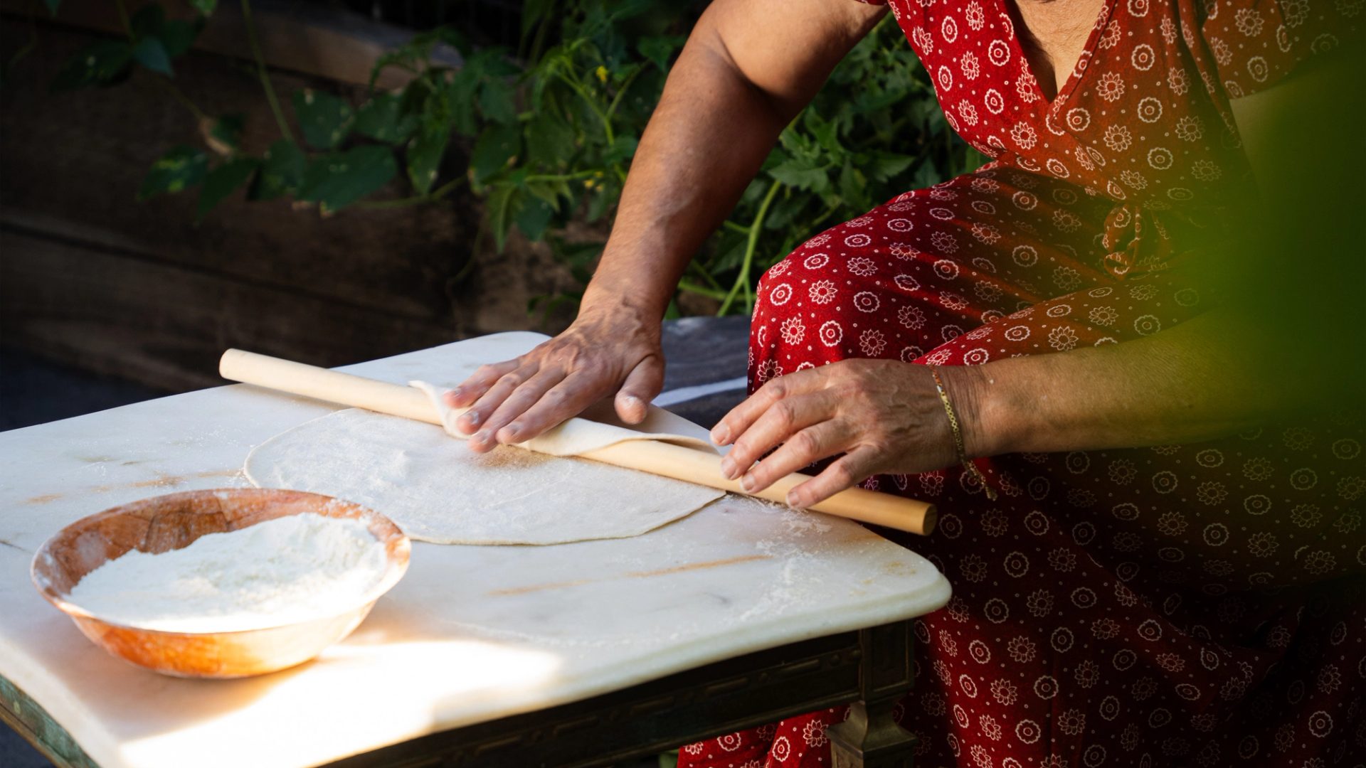 Siham is wearing a red patterned dress and is rolling out dough on a marble slab with a wooden rolling pin. The dough is being shaped into a large, thin circle. She’s rolling it out in her backyard, emphasising a natural and relaxed cooking environment.