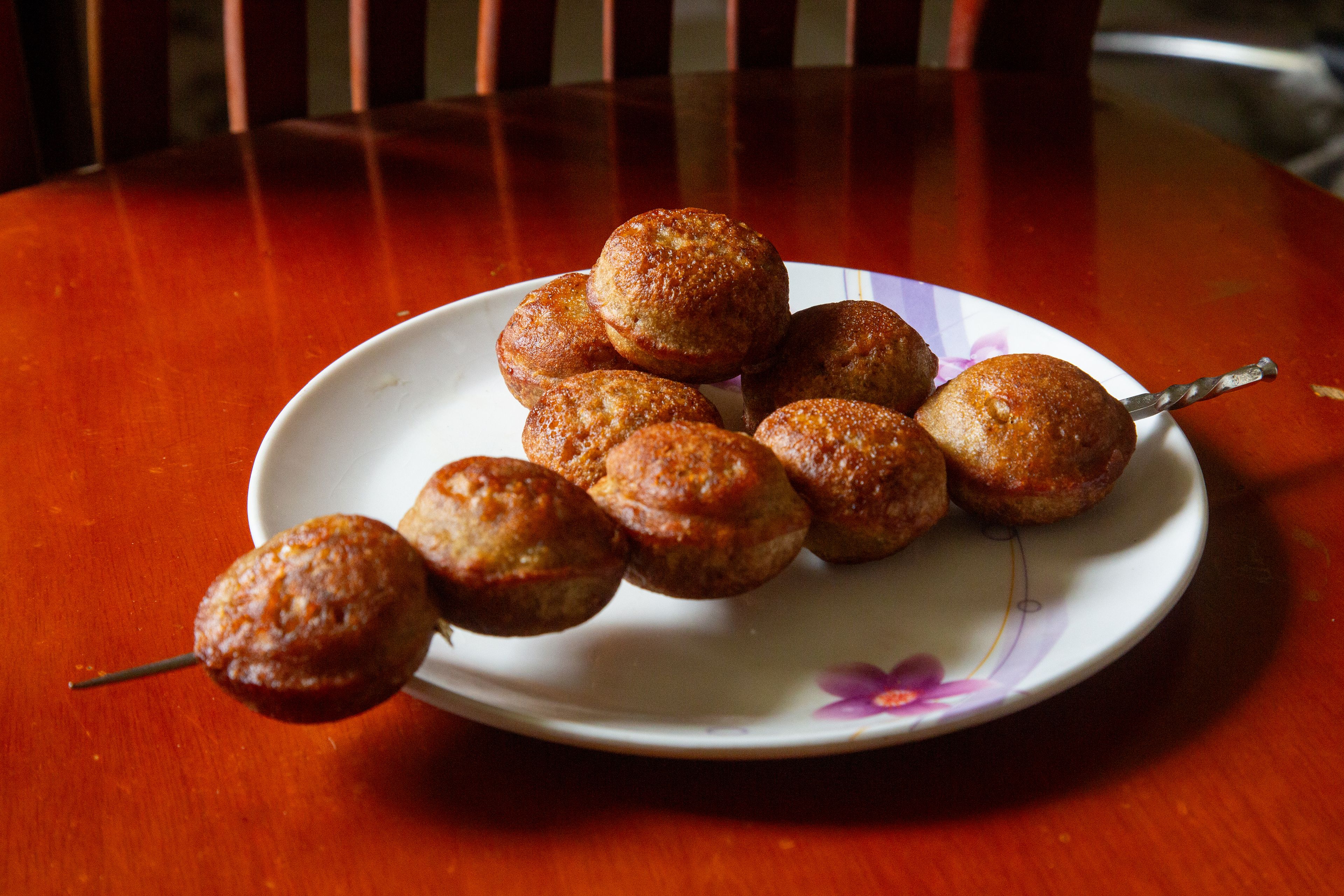 Nine small round pancake-like cooked dough balls on a white plate, five on a silver skewer.