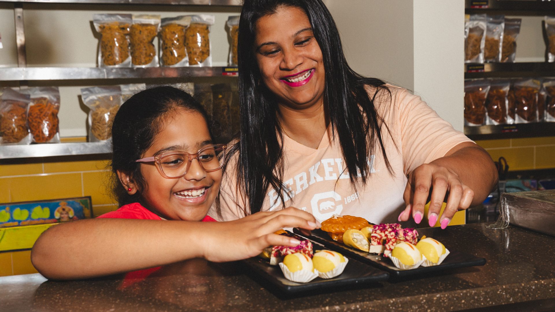 Younger figure and older figure reach for sweets atop counter.