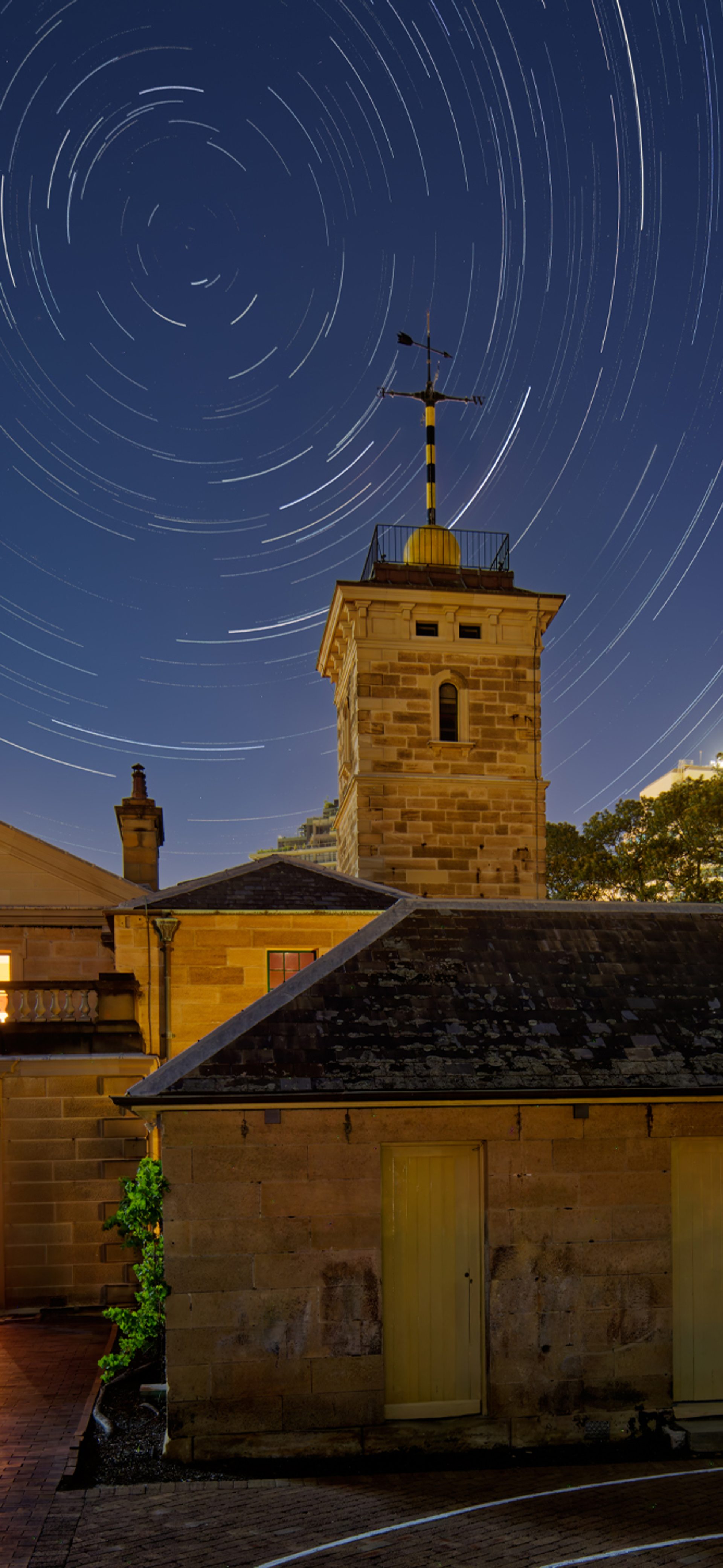 A photograph of an old yellow brick building, the Sydney Observatory, with tall city buildings and starry night sky in the background.