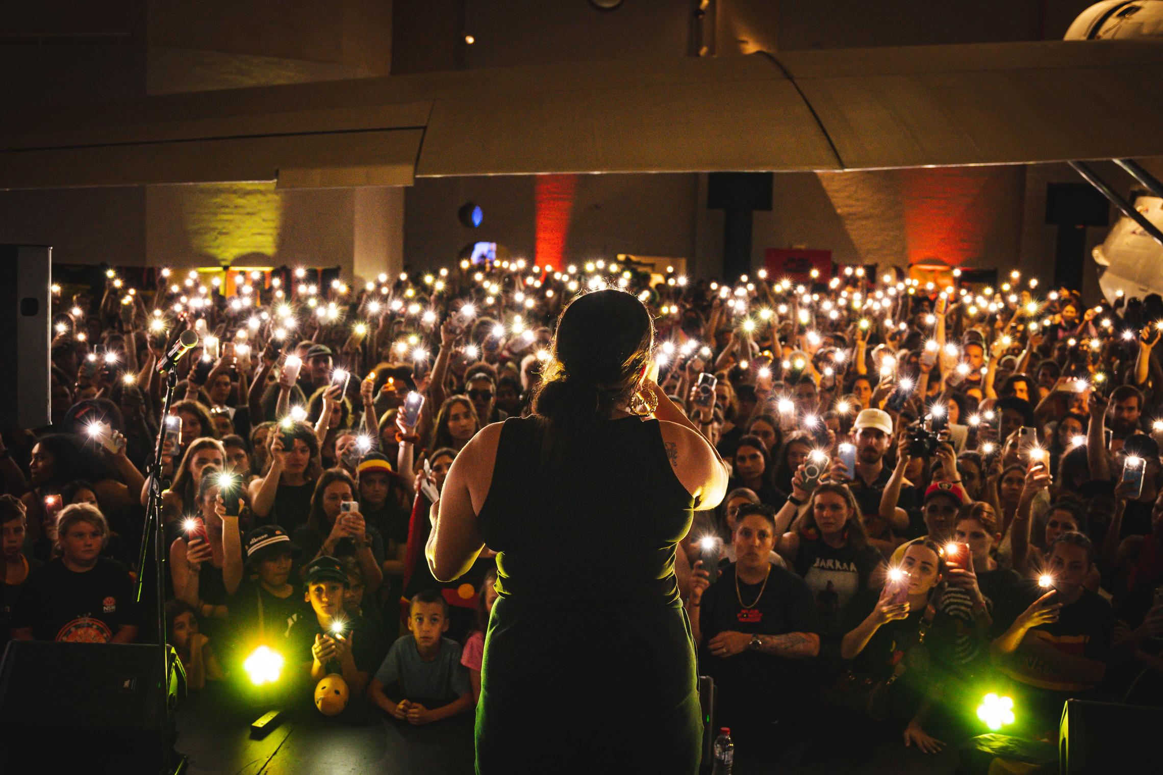 A woman performs in front of an audience who are holding their phone lights in the air.