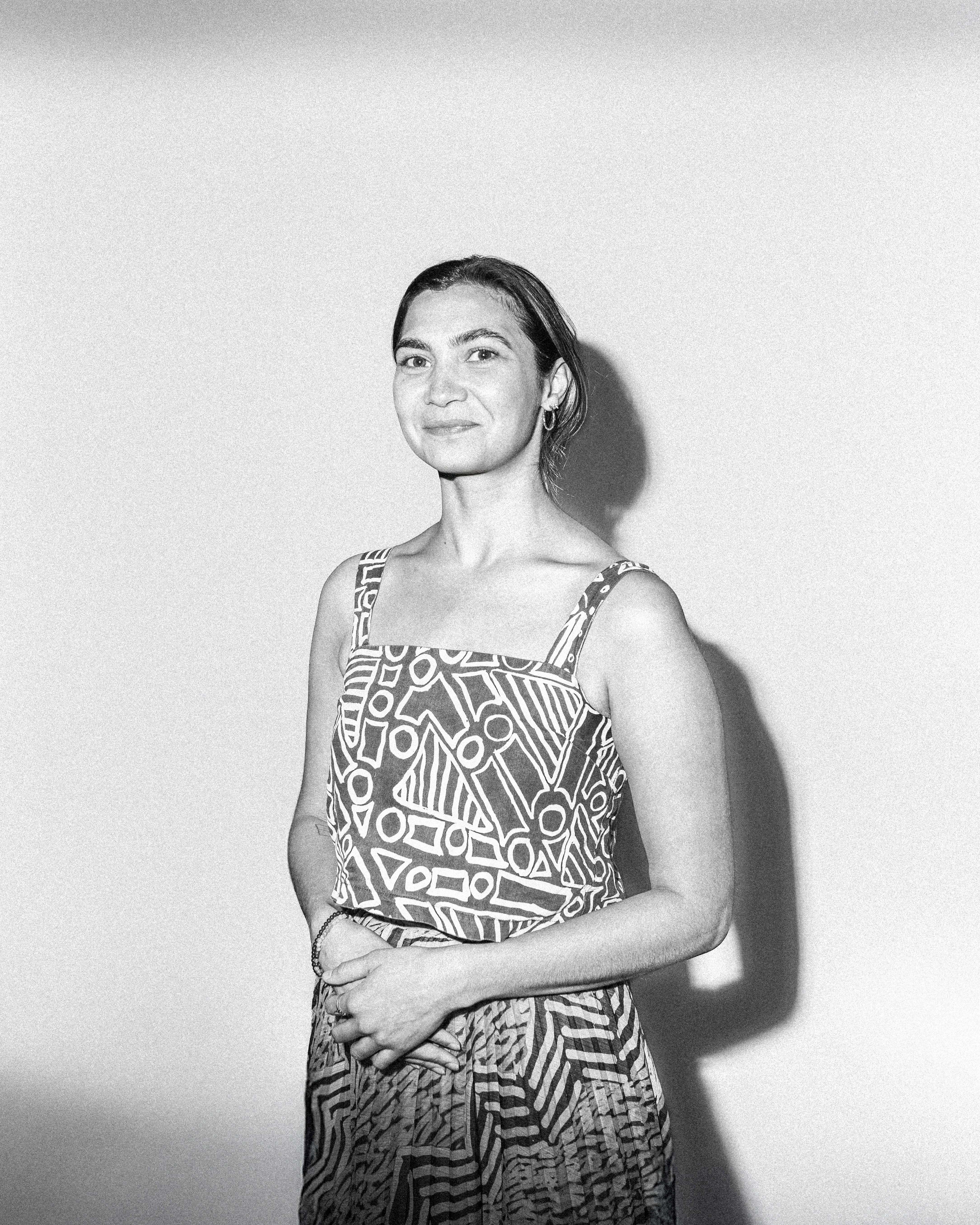 A woman with short hair wearing a strap sleeve shirt with intricate patterns (Rachael Hocking) stands against a white background, with the cameras flash casting a shadow. They are facing the camera with their hands are held together at her waist.