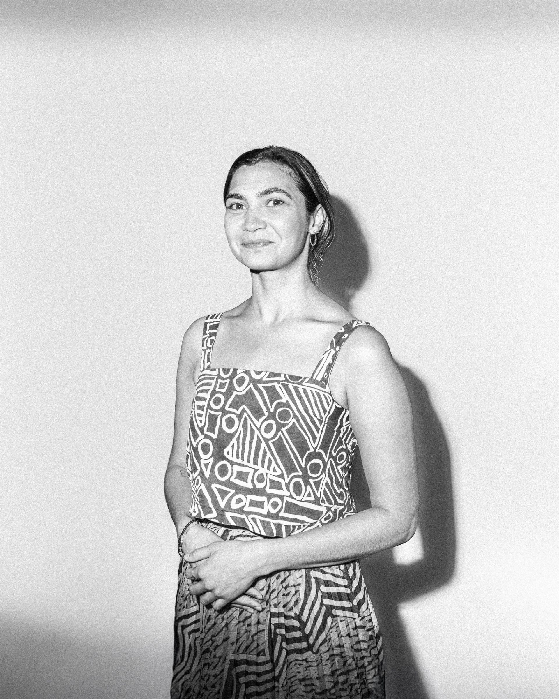 A woman with short hair wearing a strap sleeve shirt with intricate patterns (Rachael Hocking) stands against a white background, with the cameras flash casting a shadow. They are facing the camera with their hands are held together at her waist.
