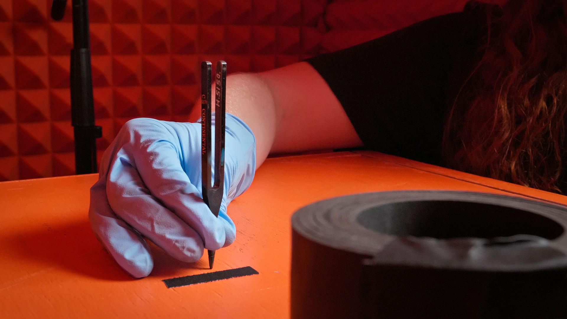 A hand in a blue plastic glove holds a metal tuning fork upright on an orange box inside an orange sound booth.