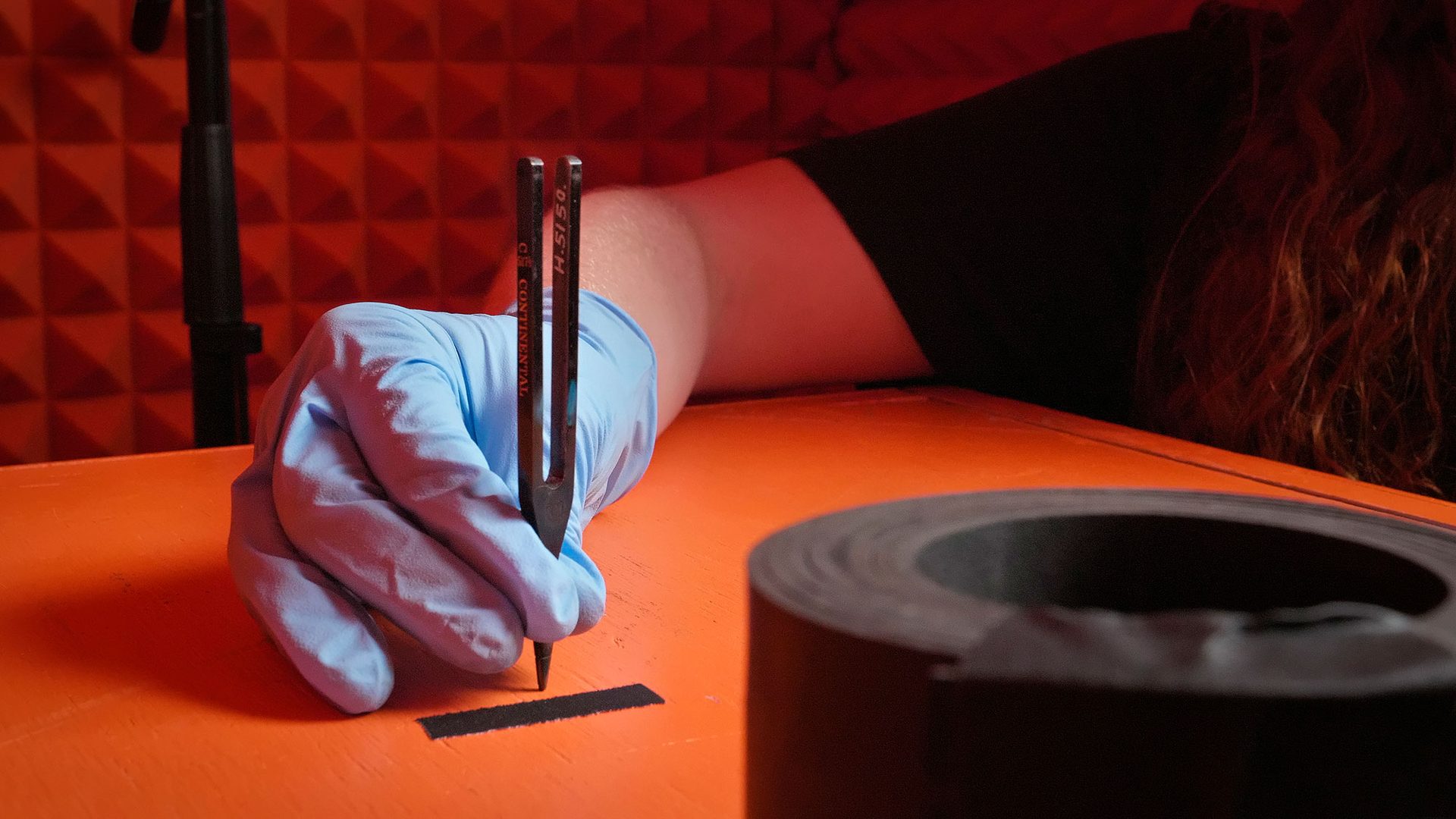 A hand in a blue plastic glove holds a metal tuning fork upright on an orange box inside an orange sound booth.