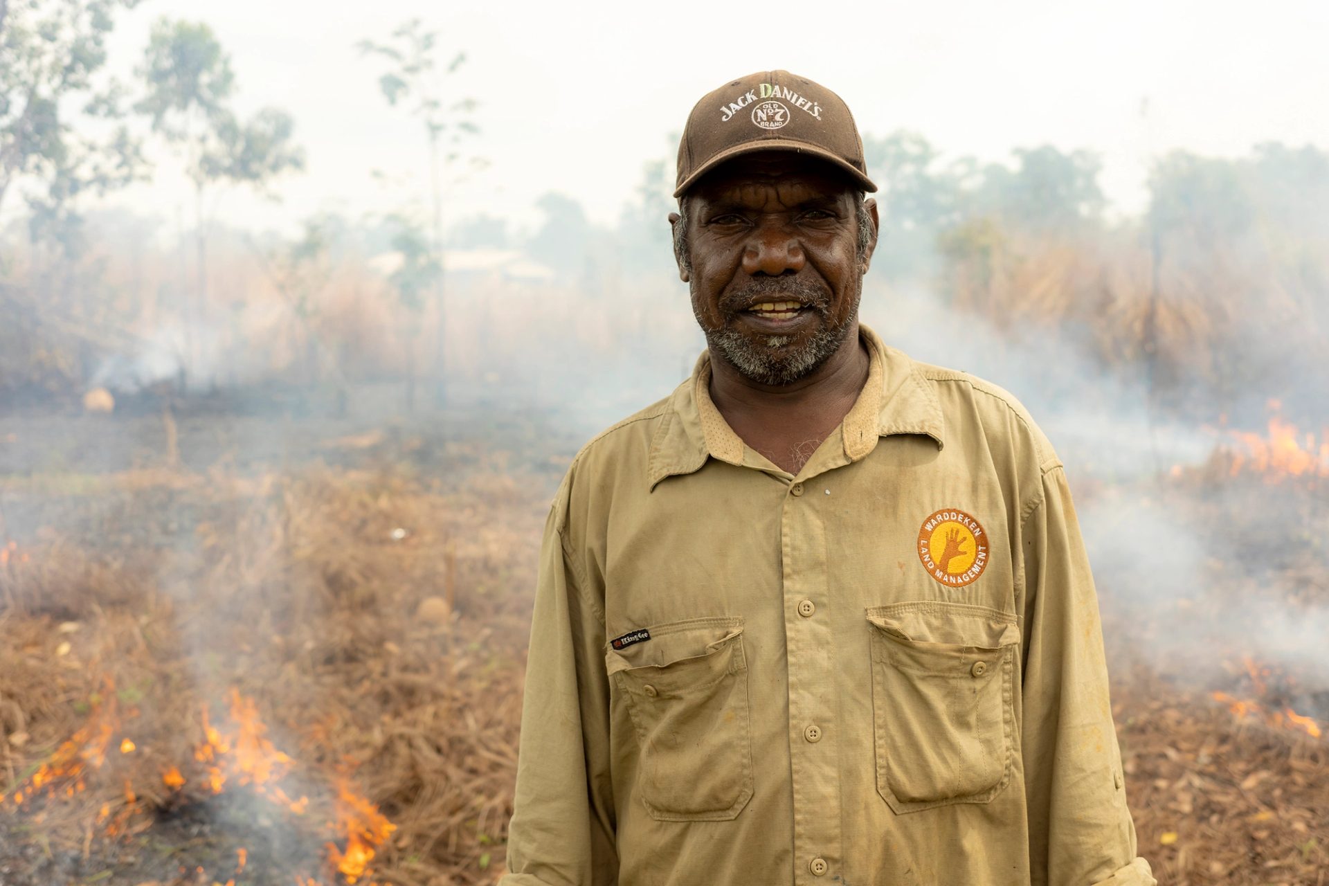 A man in a ranger uniform and a cap stands in front of a smouldering field. He is smiling at the camera.
