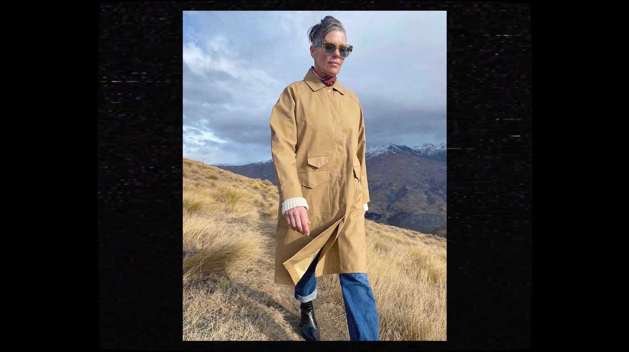 Karen Walker wearing sunglasses, a beige trench coat and jeans walking on a hill.