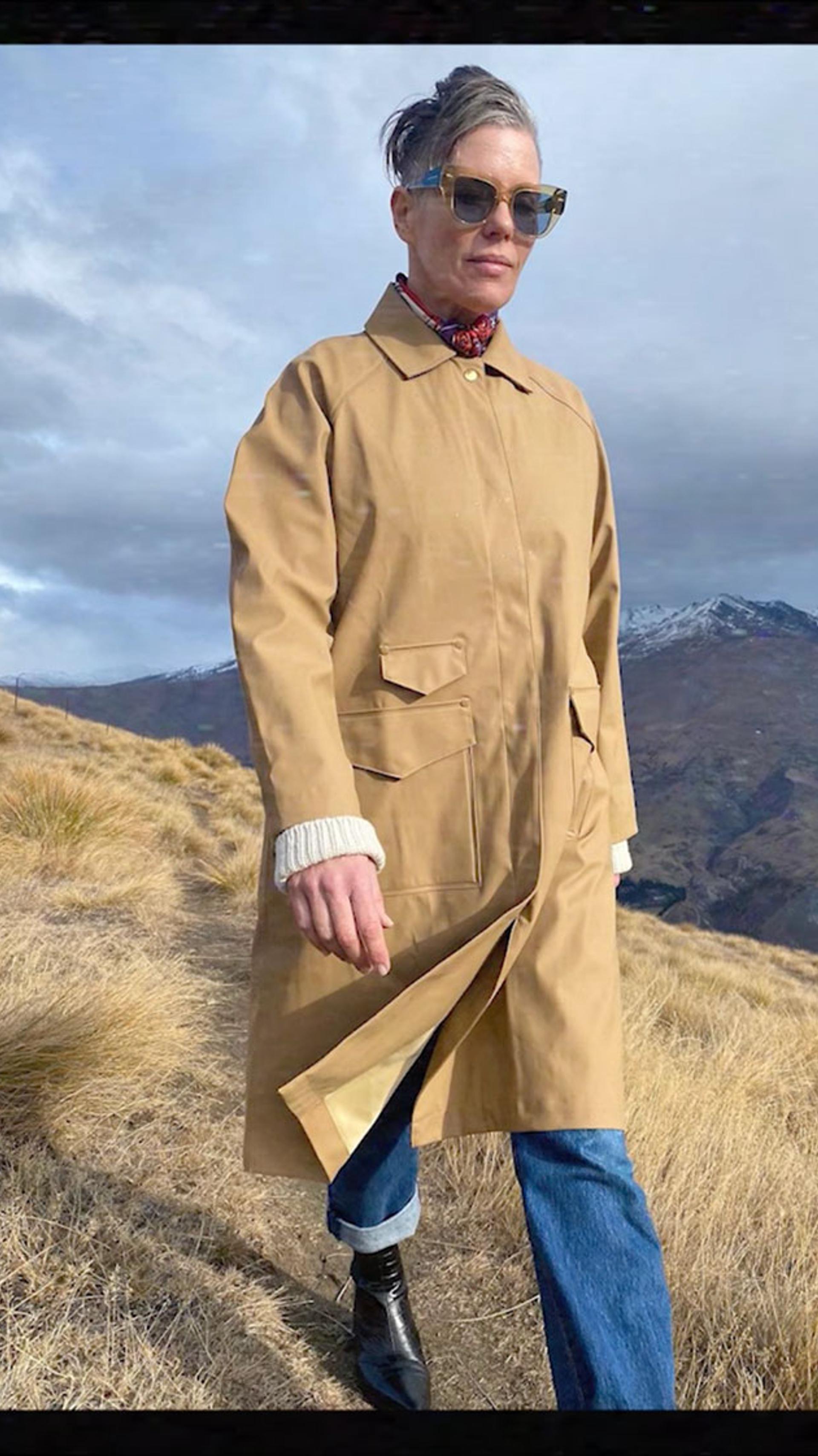 Karen Walker wearing sunglasses, a beige trench coat and jeans walking on a hill.