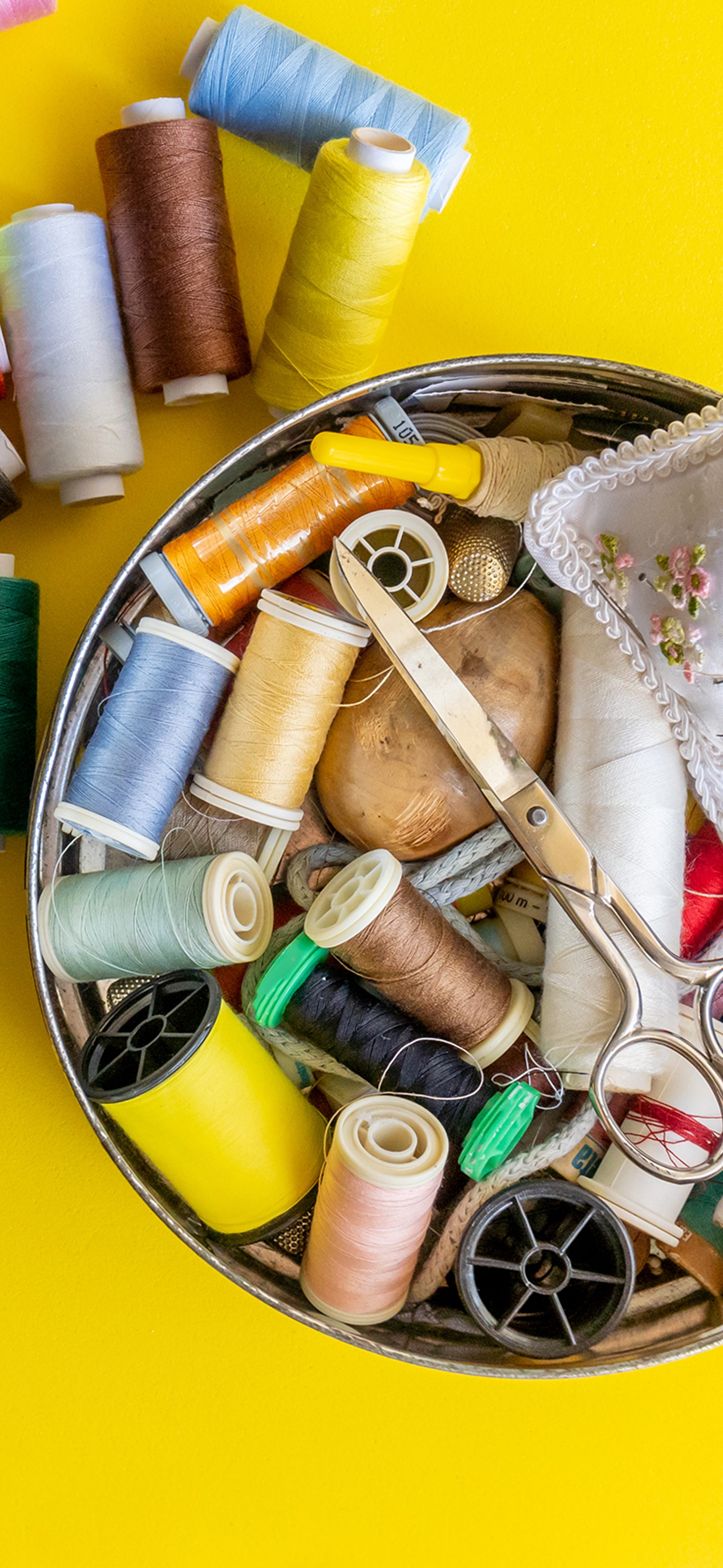 Yellow table with a overflowing tin of sewing threads, including a pair of scissors and a pin cushion.