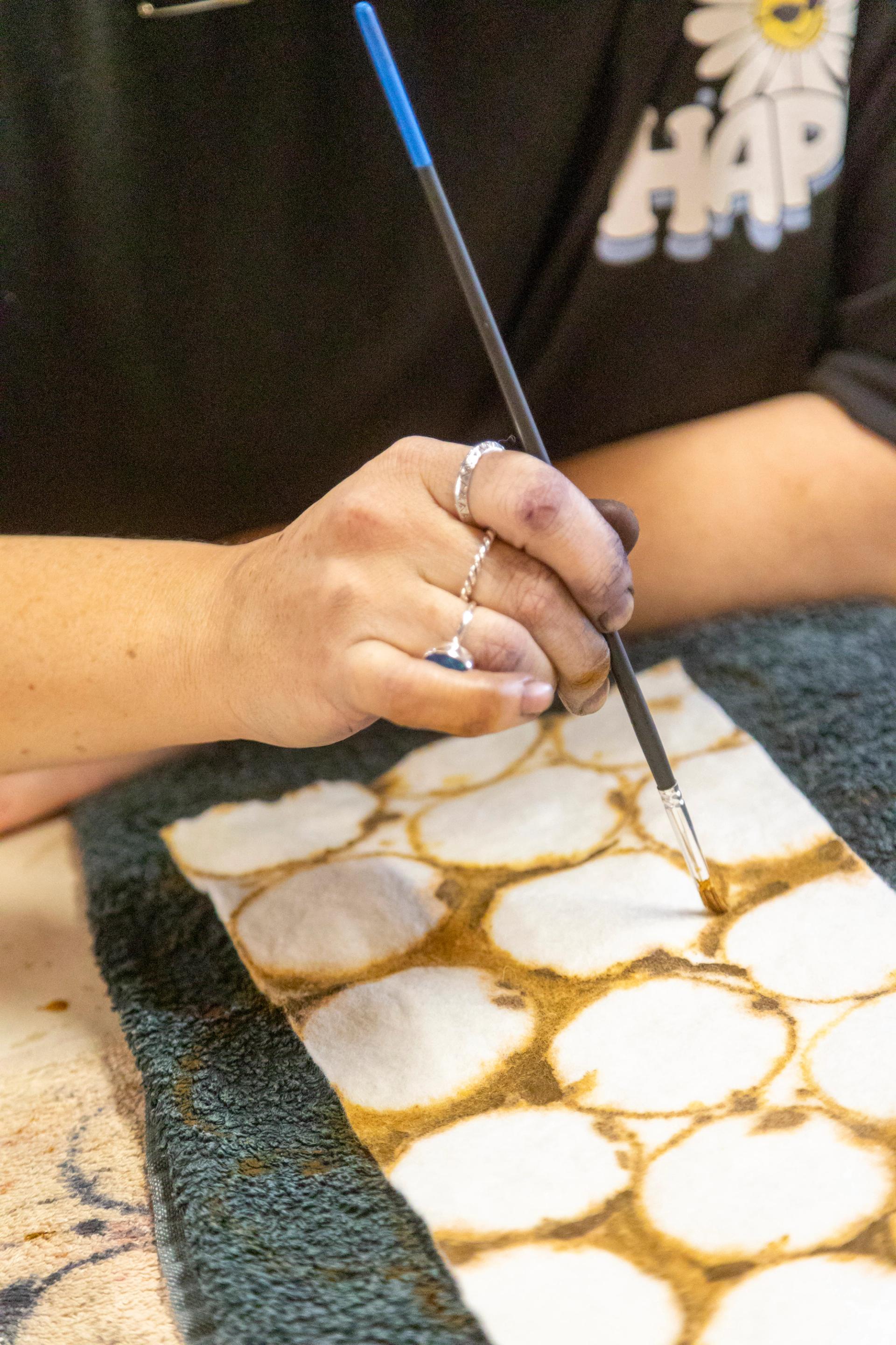 A close-up shot of a hand holding a thin paint brush painting a circular pattern on a piece of textile.