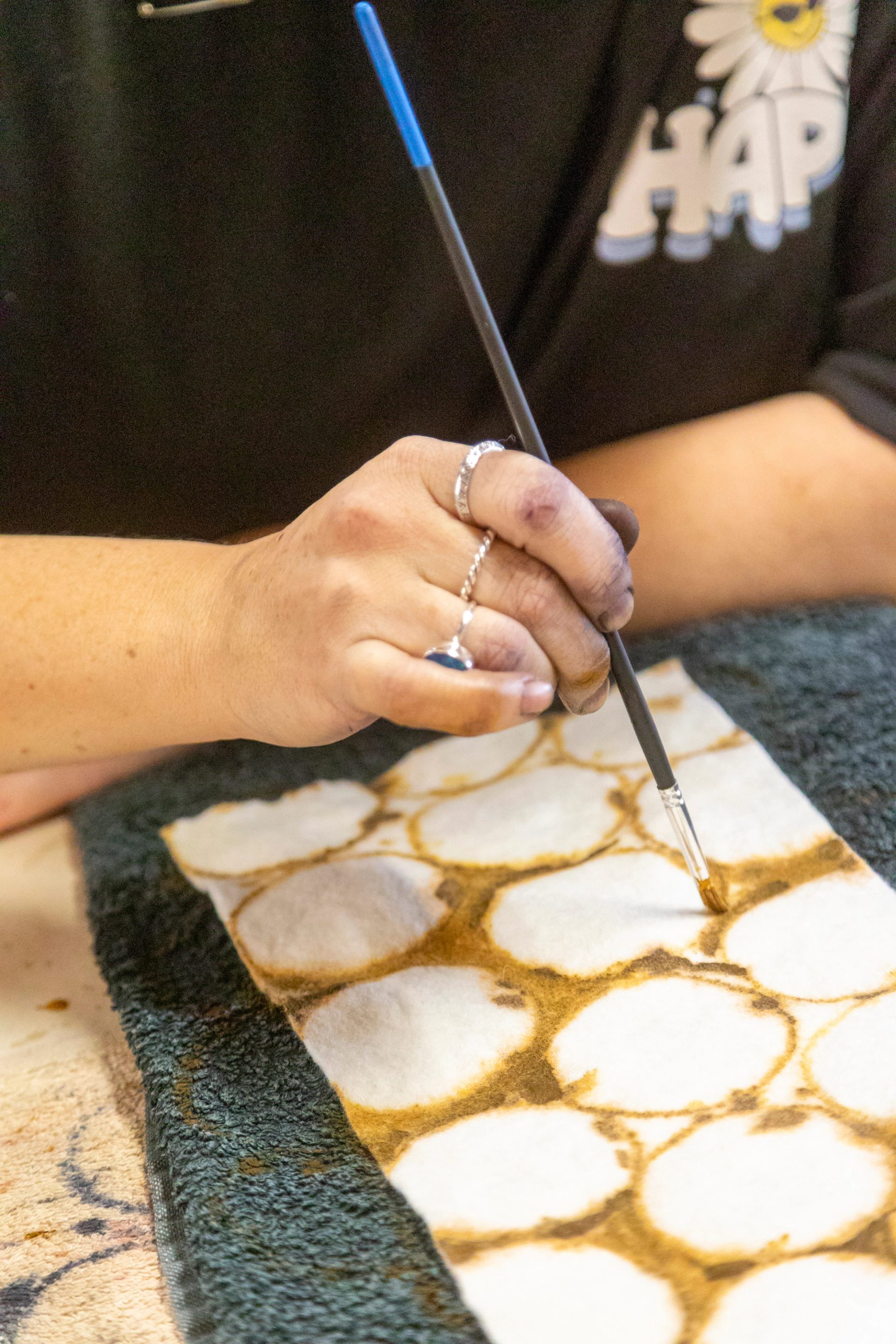 A close-up shot of a hand holding a thin paint brush painting a circular pattern on a piece of textile.