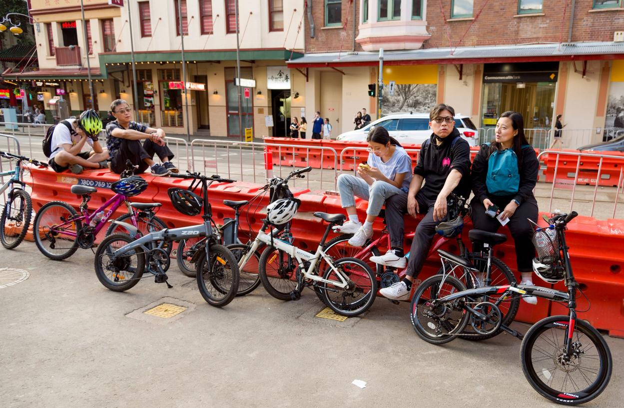 Four people sit on an orange bollard behind seven bicycles. One person is on their phone, another has their head in their hands.