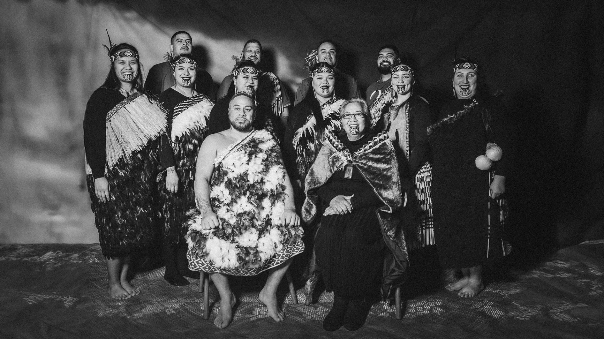 A black and white group portrait of the kapa haka group, Te Raranga Whānui. Seated at the front are Laurie Sarich, Neta Sarich. Behind them are Casey Fenton, Moana McLeod, Ashley Simon, Marei Peters, Hope Harris, Tiana Tiakiwai. At the back are Conway McLeod, Rata McGregor Jnr, Josh Simon, Royal Kingi Pita.