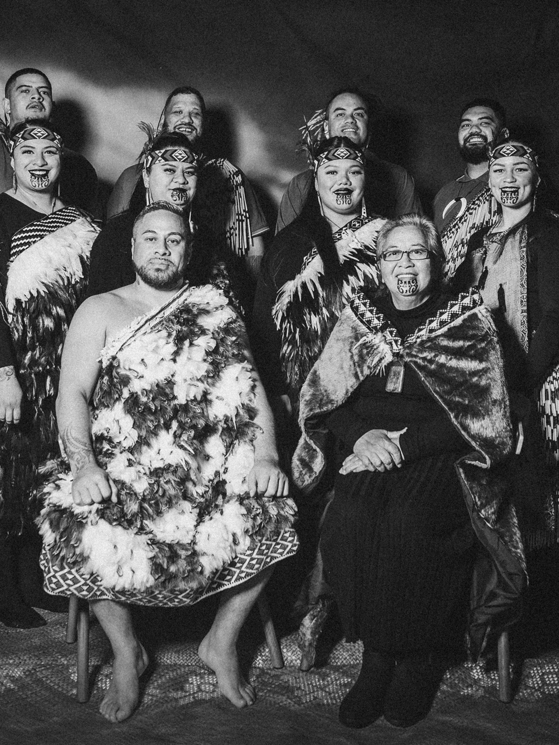 A black and white group portrait of the kapa haka group, Te Raranga Whānui. Seated at the front are Laurie Sarich, Neta Sarich. Behind them are Casey Fenton, Moana McLeod, Ashley Simon, Marei Peters, Hope Harris, Tiana Tiakiwai. At the back are Conway McLeod, Rata McGregor Jnr, Josh Simon, Royal Kingi Pita.