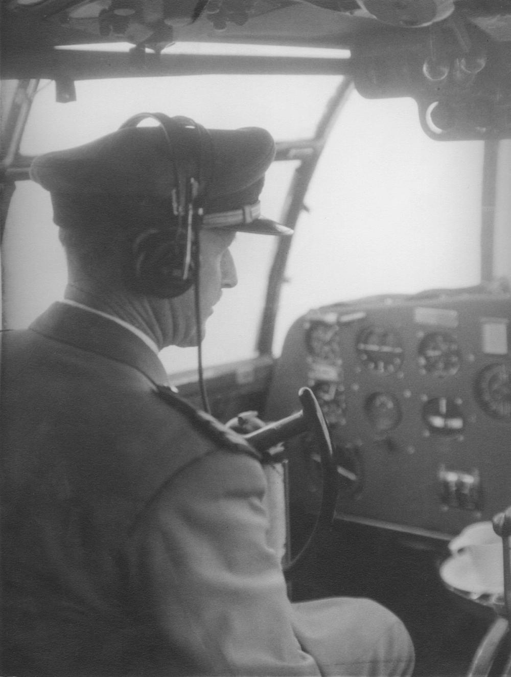Pilot in uniform sits in a cockpit of a plane wearing headphones. The photograph is black and white.