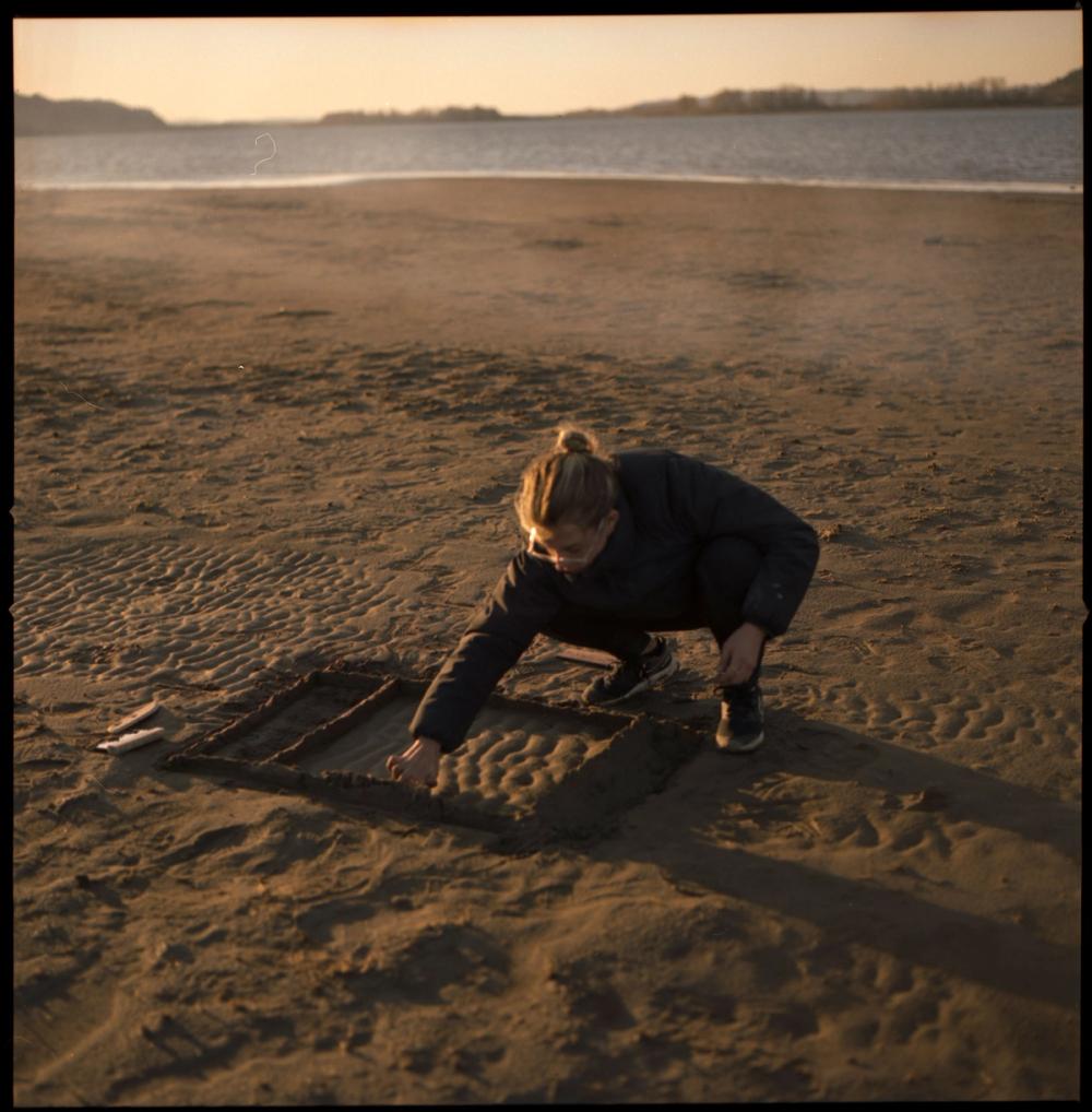 Marlo lyda crouched down on a beach working on a frame that is placed flat on the sand.