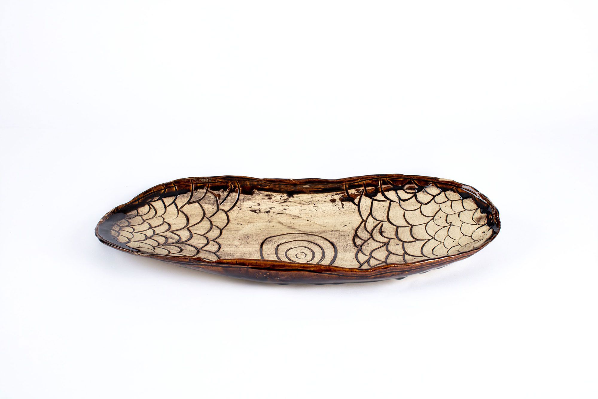 Hand formed ceramic platter shaped like a coolamon. The design in the bowl features incised concentric circles in the centre and a number of arc designs at both ends. Dark brown iron oxide glaze around edge of dish and in grooves of design.
