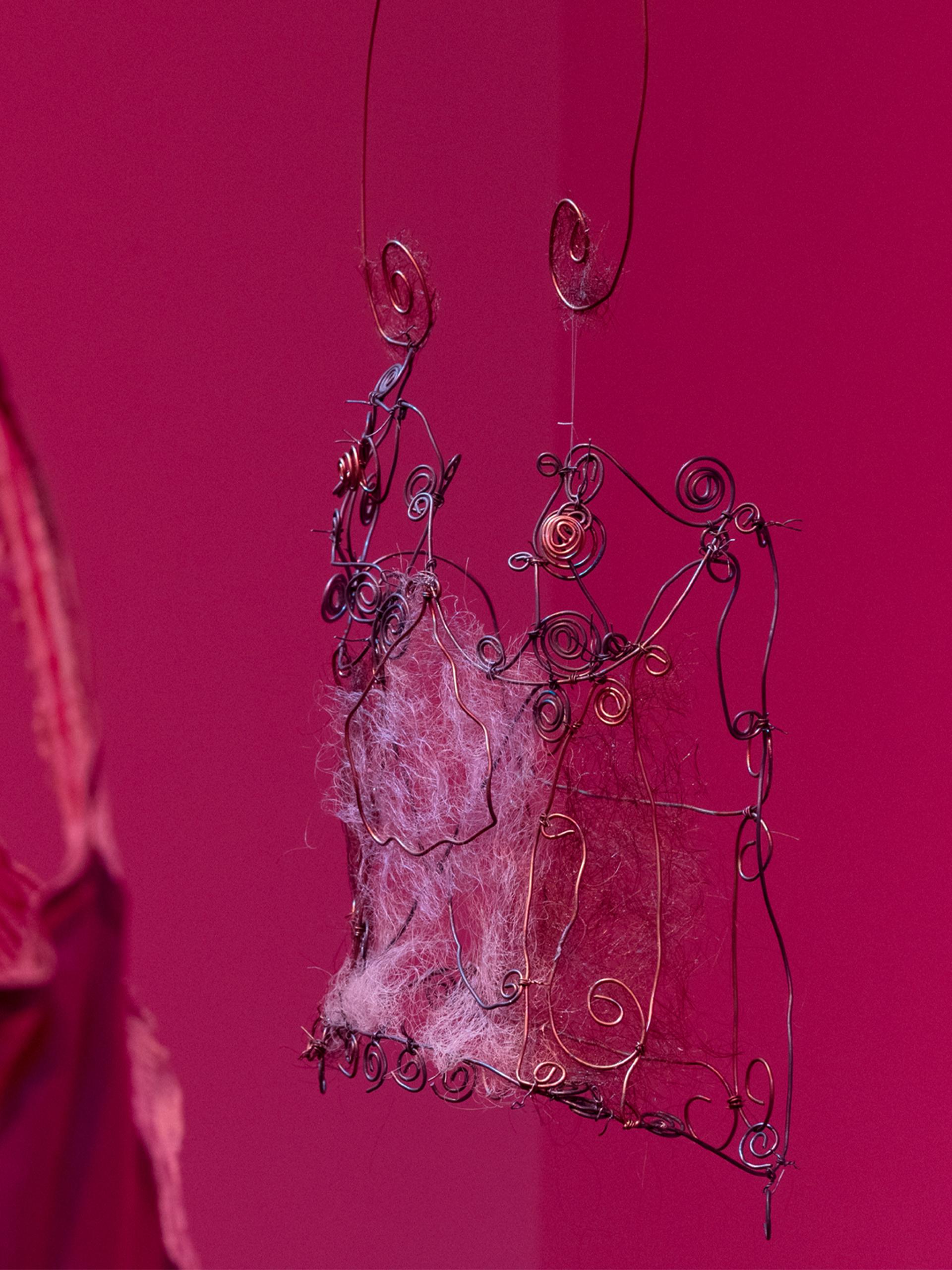A close-up of We Are Studios artist Rebecca Gentz Sciroli’s In the shape of her absence, honouring women lost to domestic violence and drawing on her lived experience as a survivor. In deep pink-red light, a delicate wire corset woven with human hair from three generations of Rebecca’s family of women hangs in focus on the right, while part of a mauve satin corset-slip appears softly out of focus on the left.