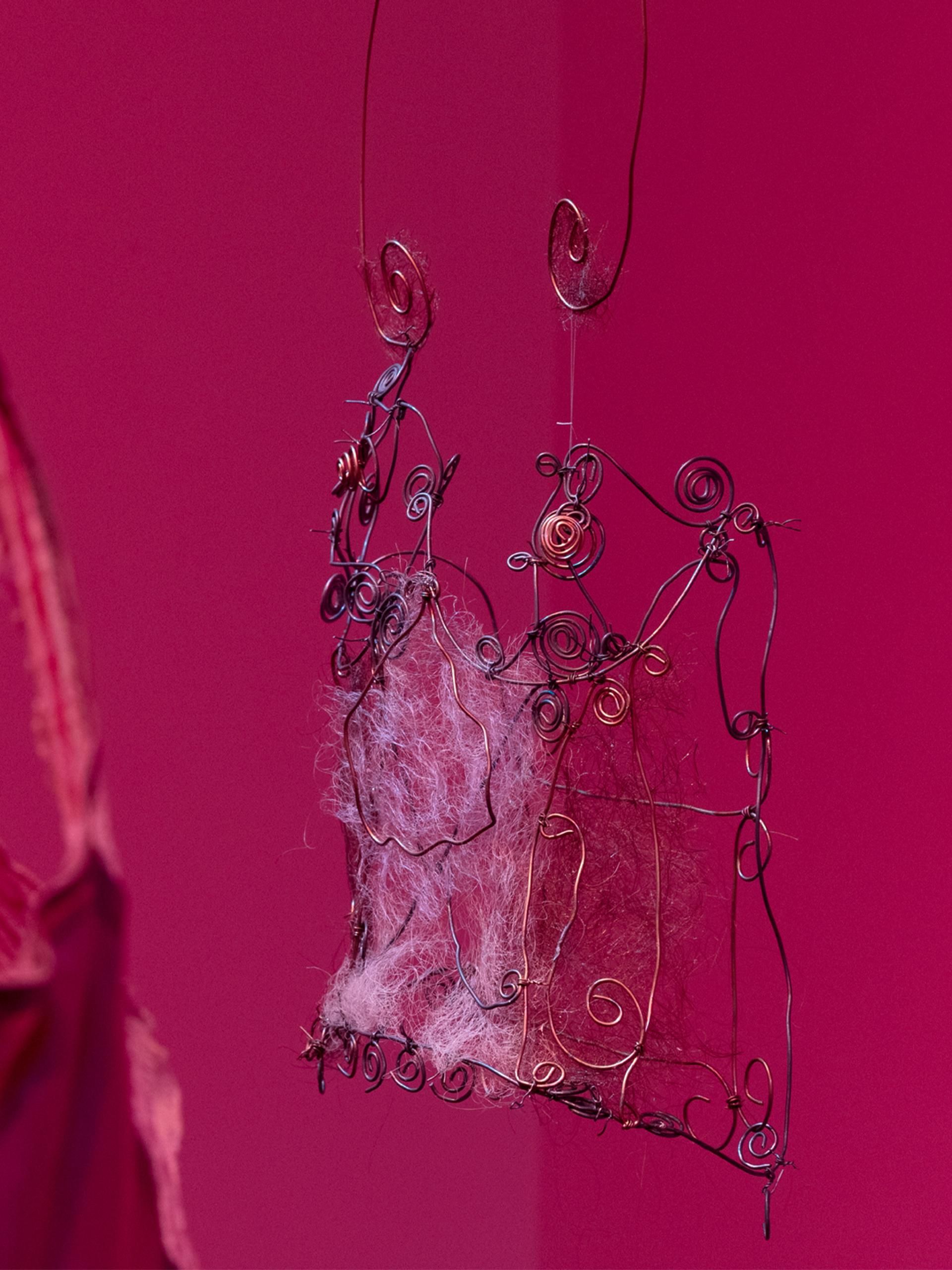A close-up of We Are Studios artist Rebecca Gentz Sciroli’s In the shape of her absence, honouring women lost to domestic violence and drawing on her lived experience as a survivor. In deep pink-red light, a delicate wire corset woven with human hair from three generations of Rebecca’s family of women hangs in focus on the right, while part of a mauve satin corset-slip appears softly out of focus on the left.