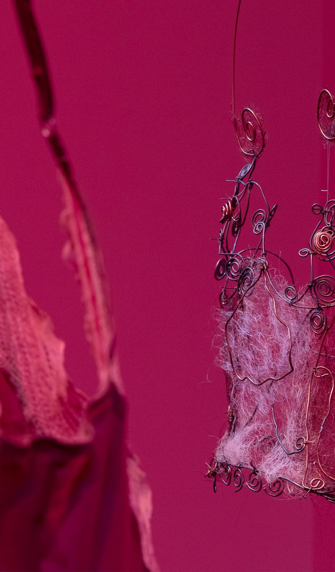 A close-up of We Are Studios artist Rebecca Gentz Sciroli’s In the shape of her absence, honouring women lost to domestic violence and drawing on her lived experience as a survivor. In deep pink-red light, a delicate wire corset woven with human hair from three generations of Rebecca’s family of women hangs in focus on the right, while part of a mauve satin corset-slip appears softly out of focus on the left.