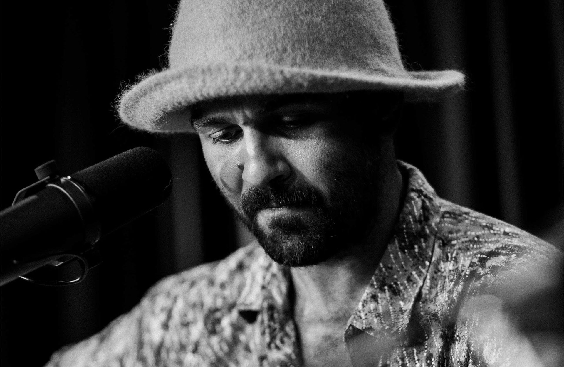 A black and white image of Joseph Douglas wearing a felt hat and patterned shirt. He is behind a microphone looking down with a concentrated face.