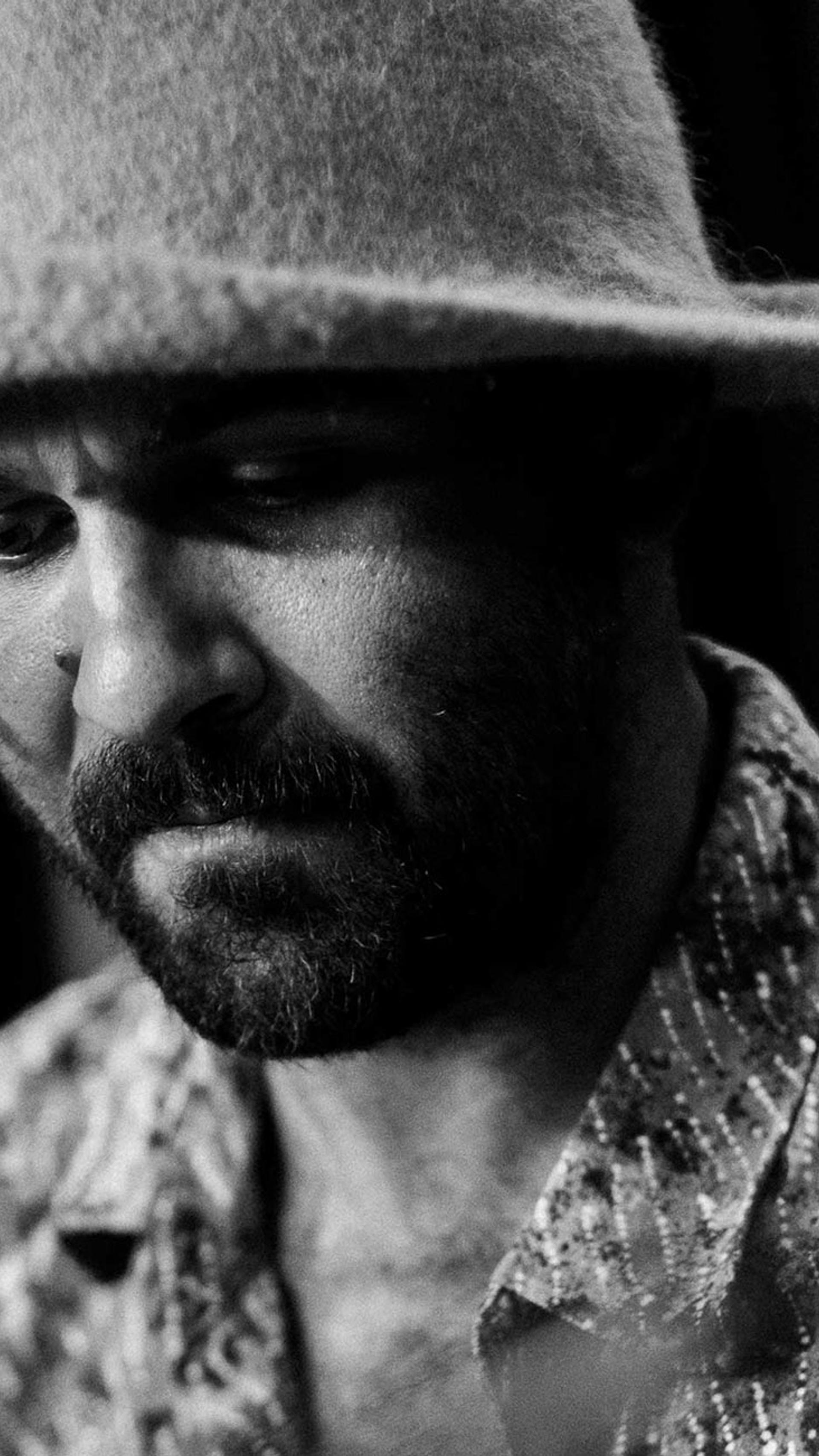 A black and white image of Joseph Douglas wearing a felt hat and patterned shirt. He is behind a microphone looking down with a concentrated face.