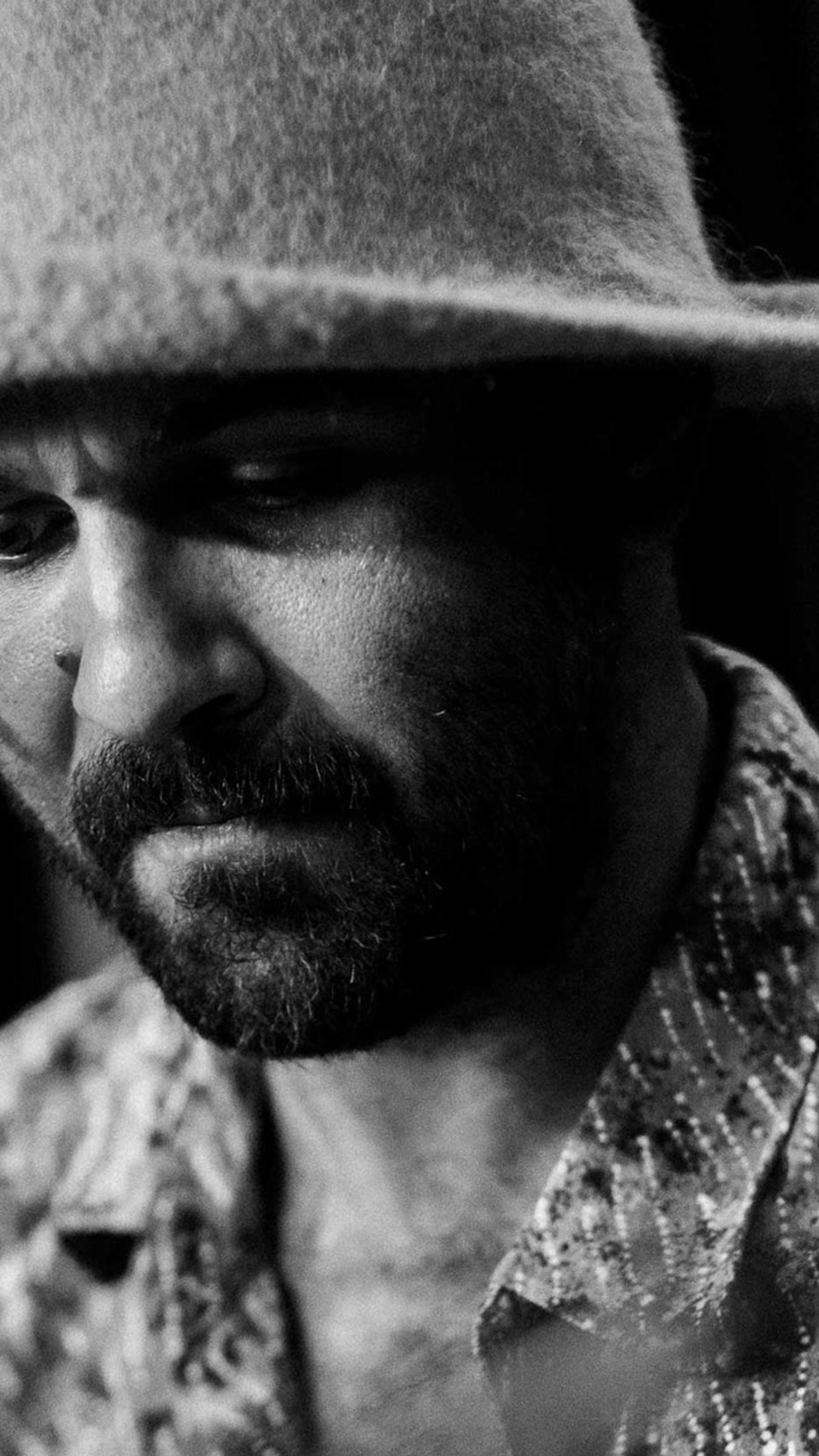 A black and white image of Joseph Douglas wearing a felt hat and patterned shirt. He is behind a microphone looking down with a concentrated face.