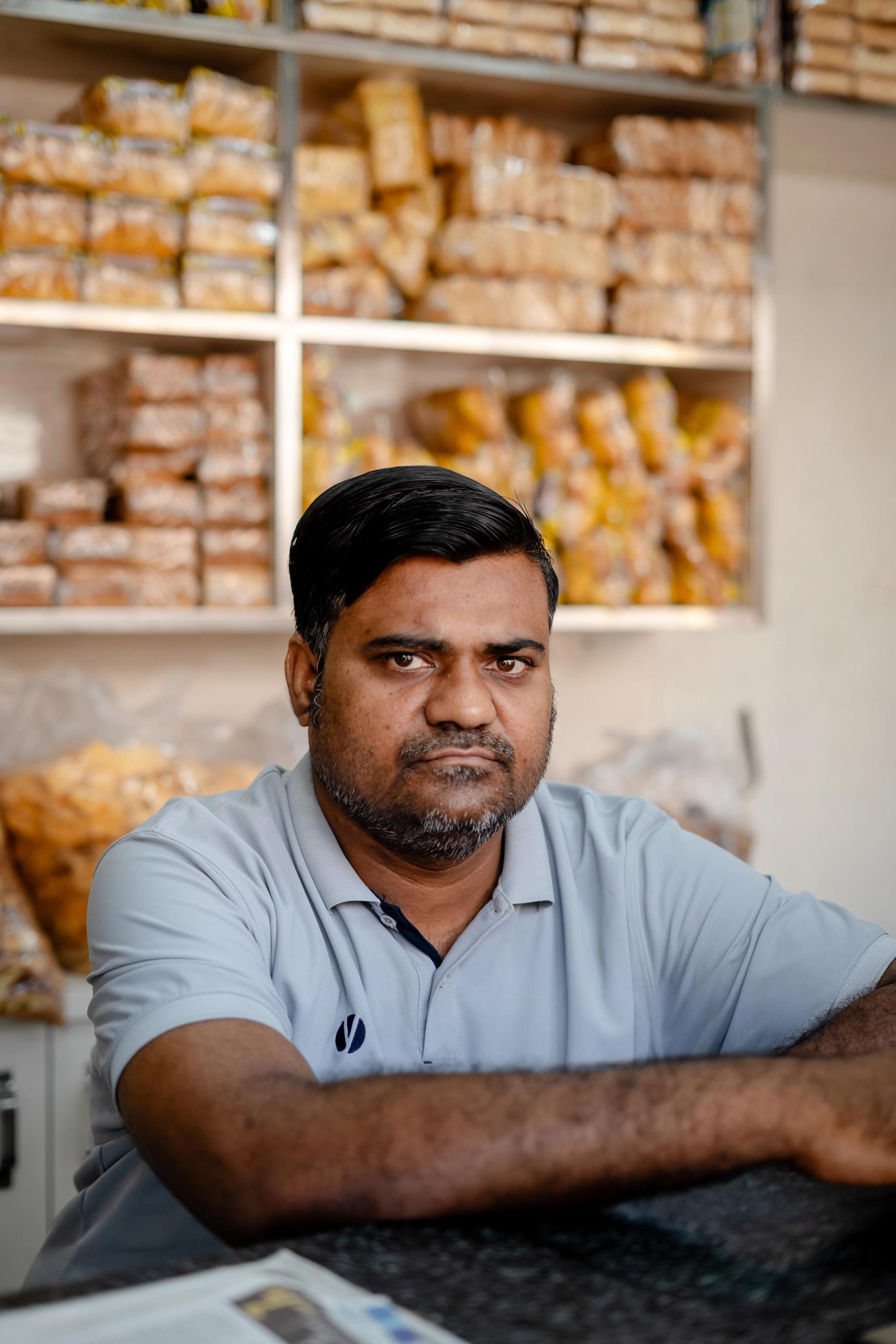 A man in a bakery, stands in front of two shelves filled with bread rolls. He wears a blue polo shirt.