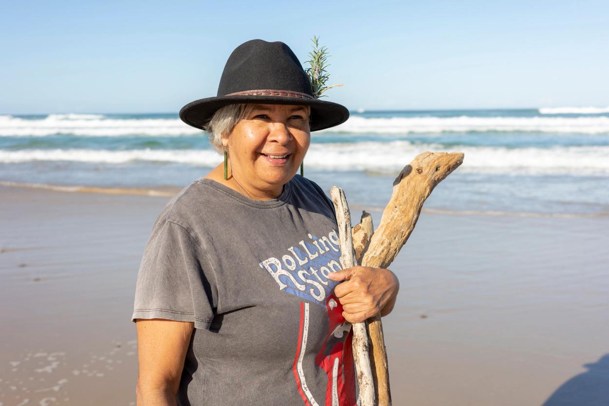 A mid shot of a woman standing on a beach and holding three pieces of driftwood in her left arm.