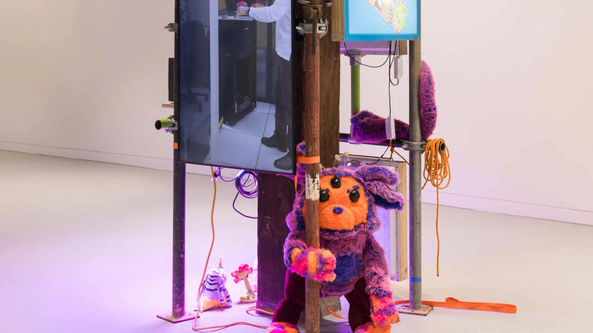 Photo of an indoor art installation that comprises scaffolding with a video screen and a soft sculpture of a creature with an orange face and purple ears.