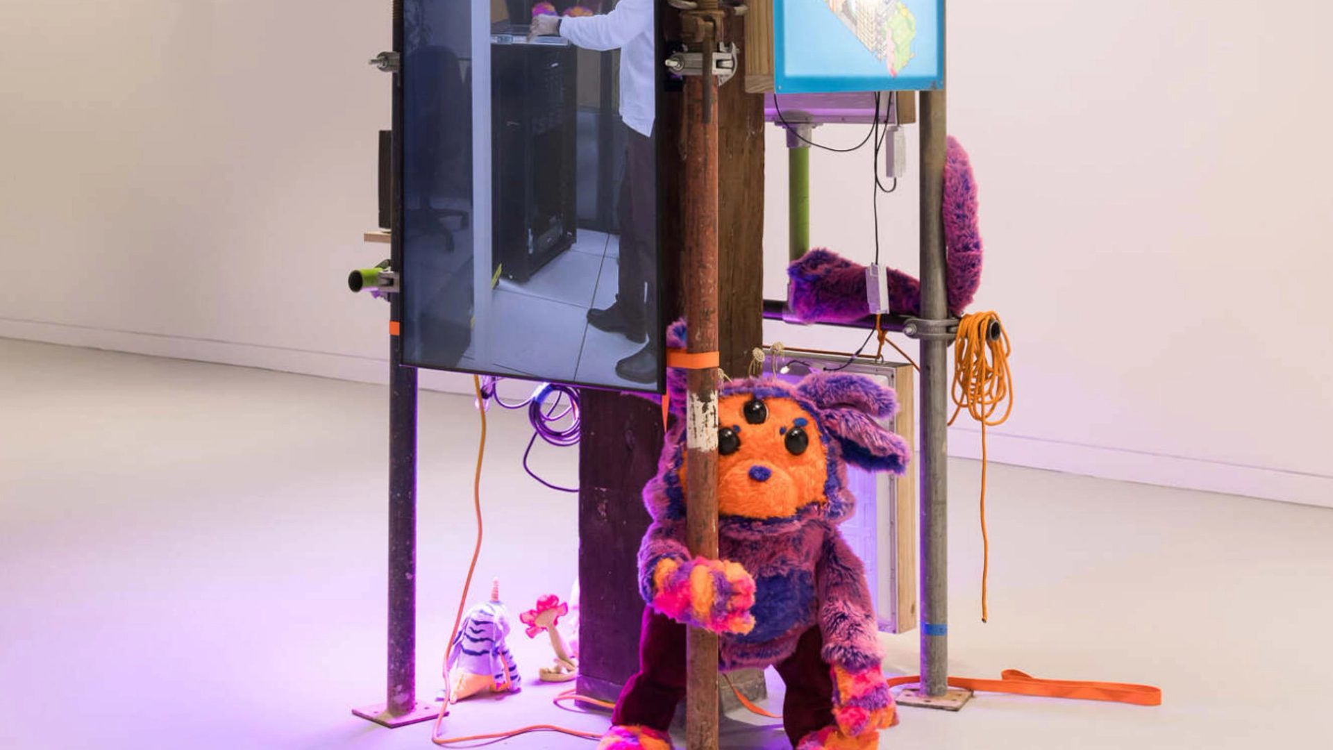 Photo of an indoor art installation that comprises scaffolding with a video screen and a soft sculpture of a creature with an orange face and purple ears.