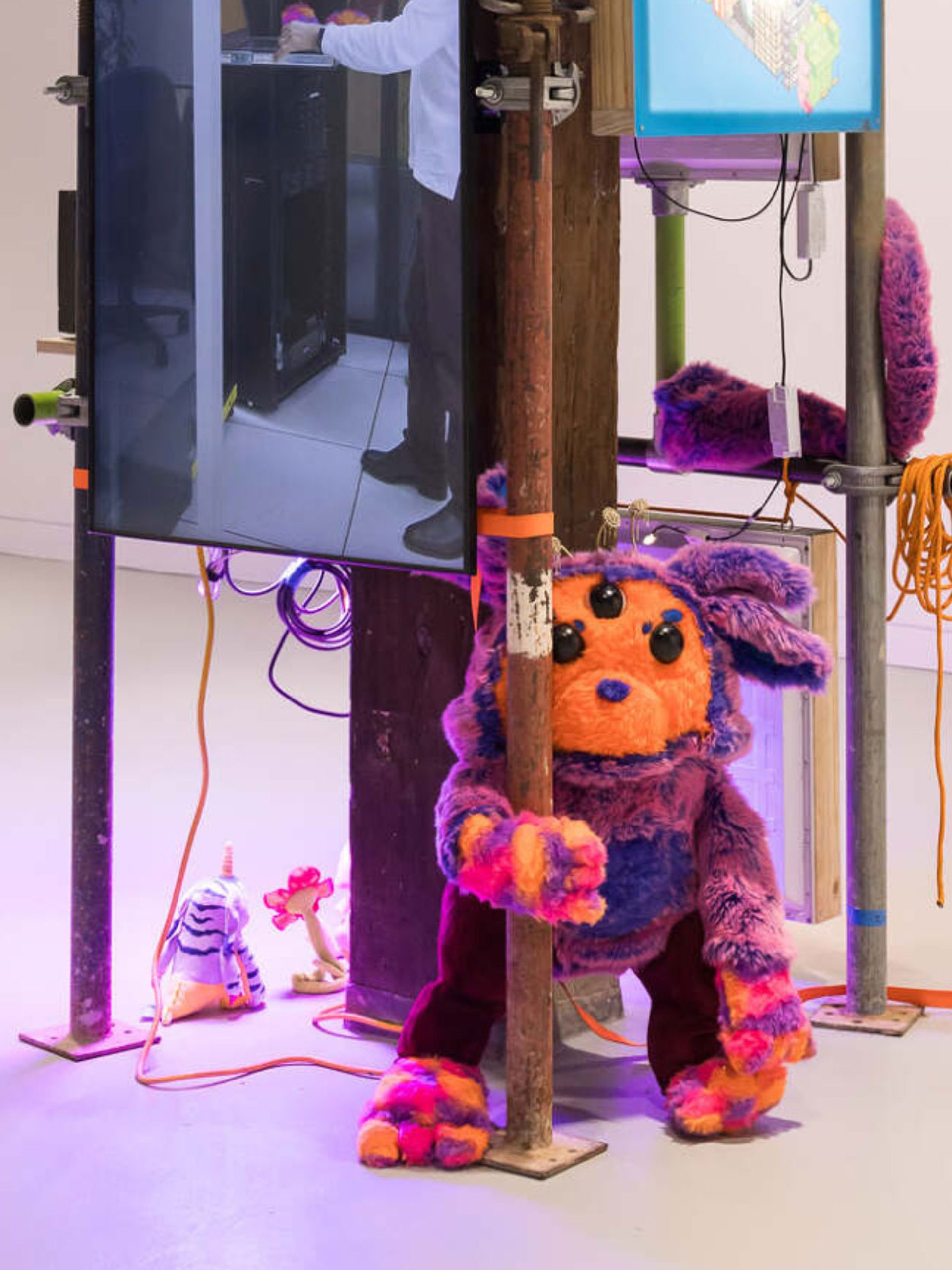 Photo of an indoor art installation that comprises scaffolding with a video screen and a soft sculpture of a creature with an orange face and purple ears.