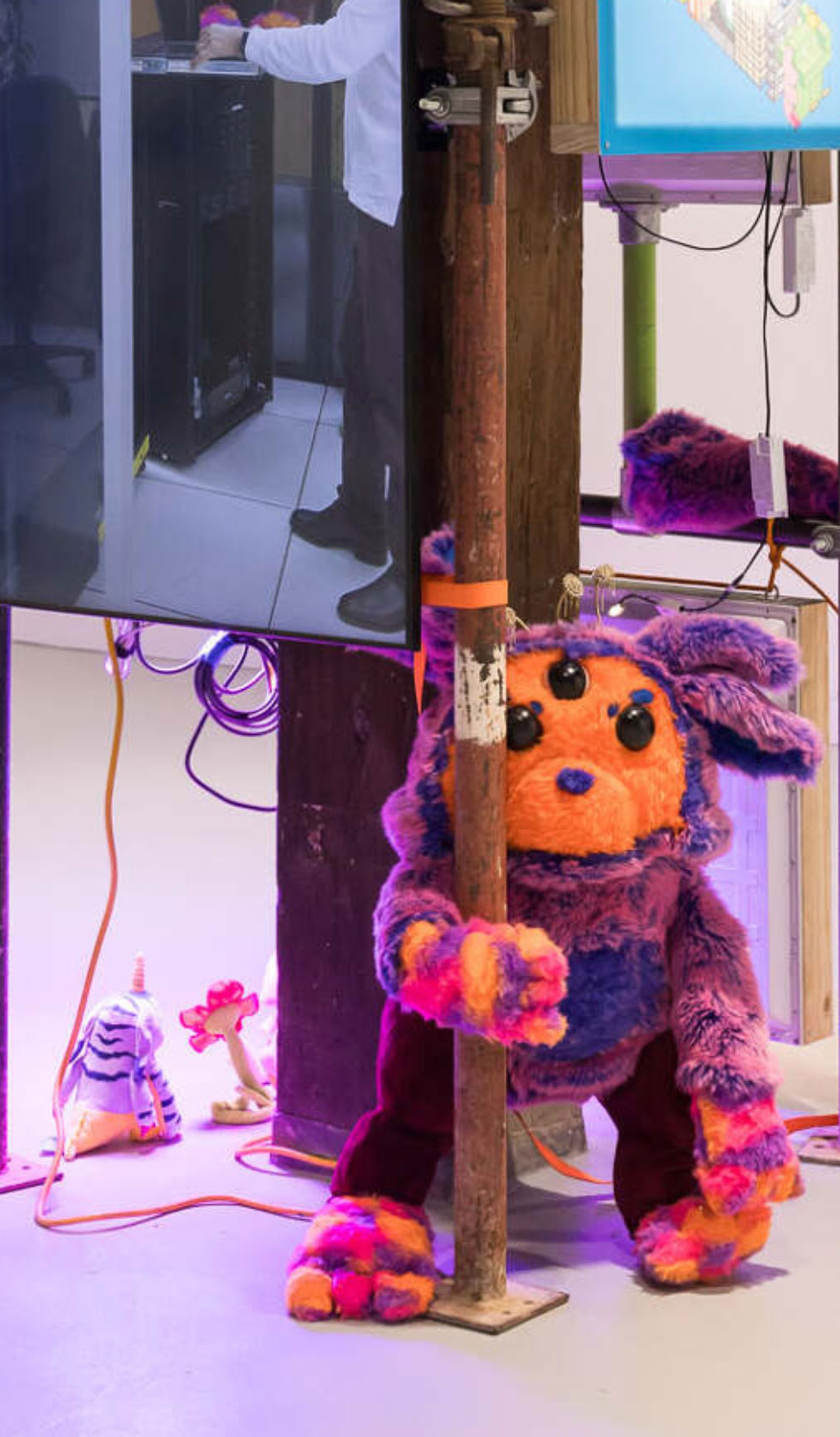 Photo of an indoor art installation that comprises scaffolding with a video screen and a soft sculpture of a creature with an orange face and purple ears.