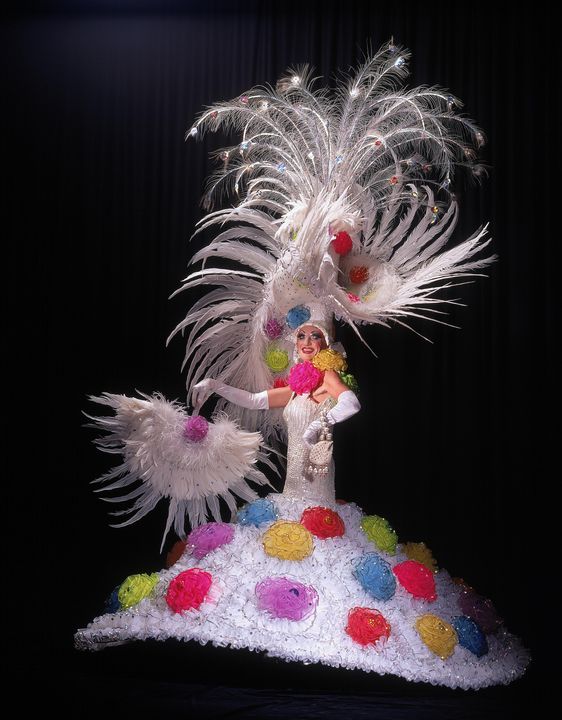 A white sleeveless, fitted sequinned dress to which is velcroed a full skirt of white tulle decorated with multicoloured tulle rosettes