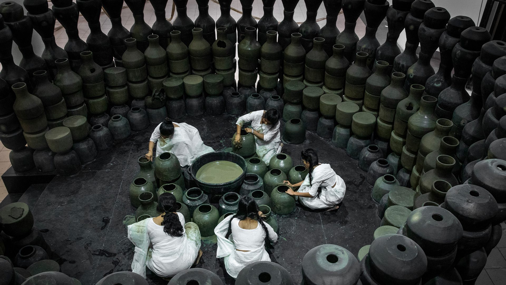 Five people dressed in white sit on the floor around black pots and surrounded by black pots.