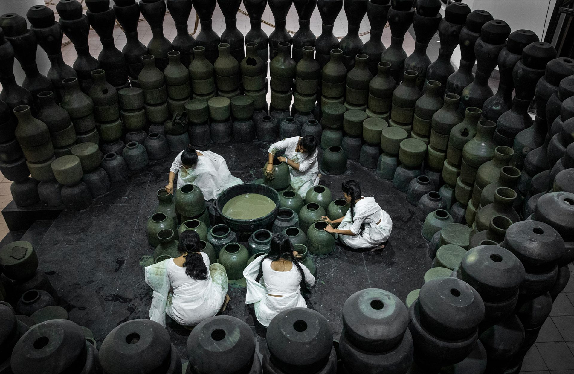 Five people dressed in white sit on the floor around black pots and surrounded by black pots.