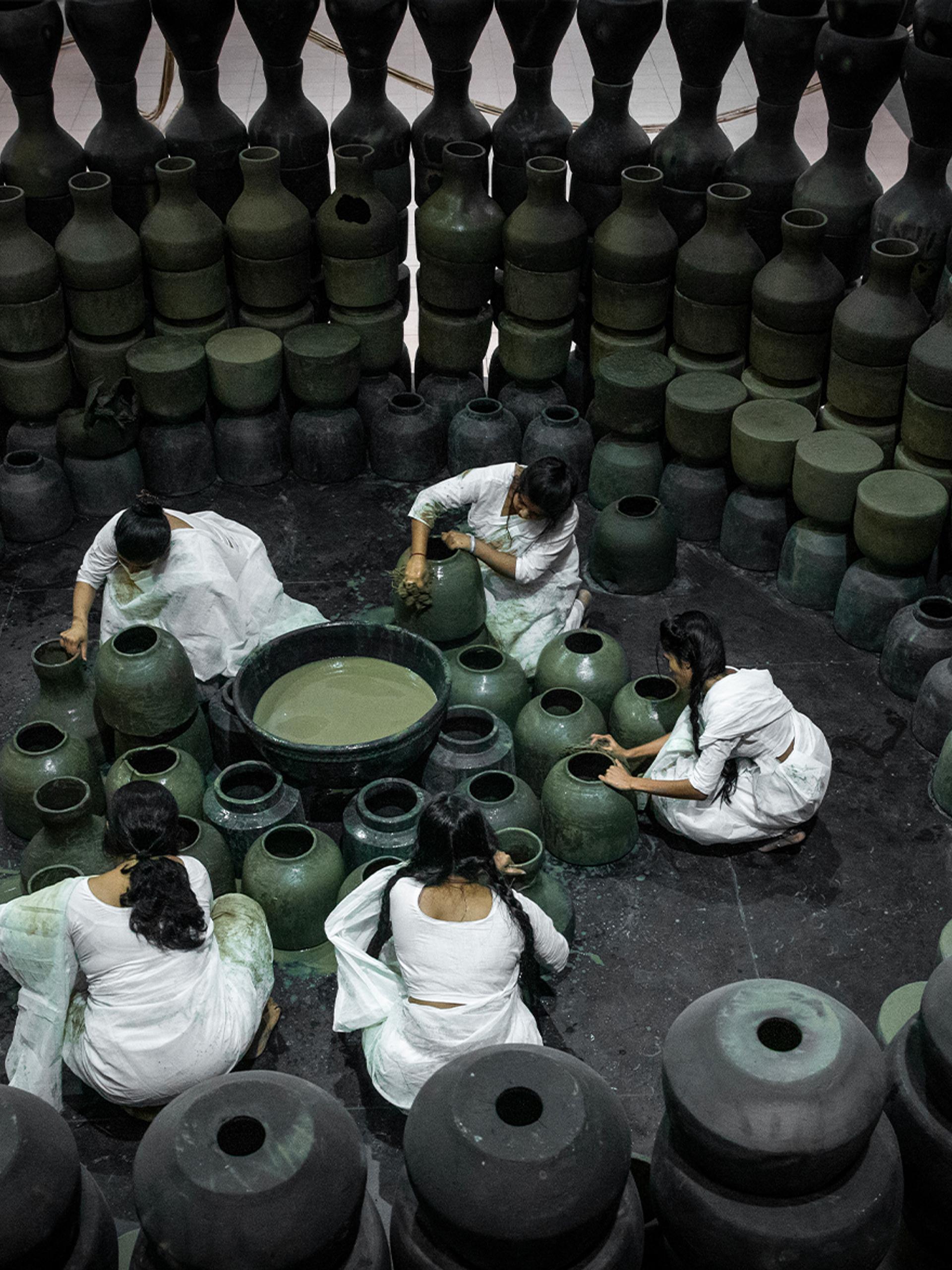Five people dressed in white sit on the floor around black pots and surrounded by black pots.