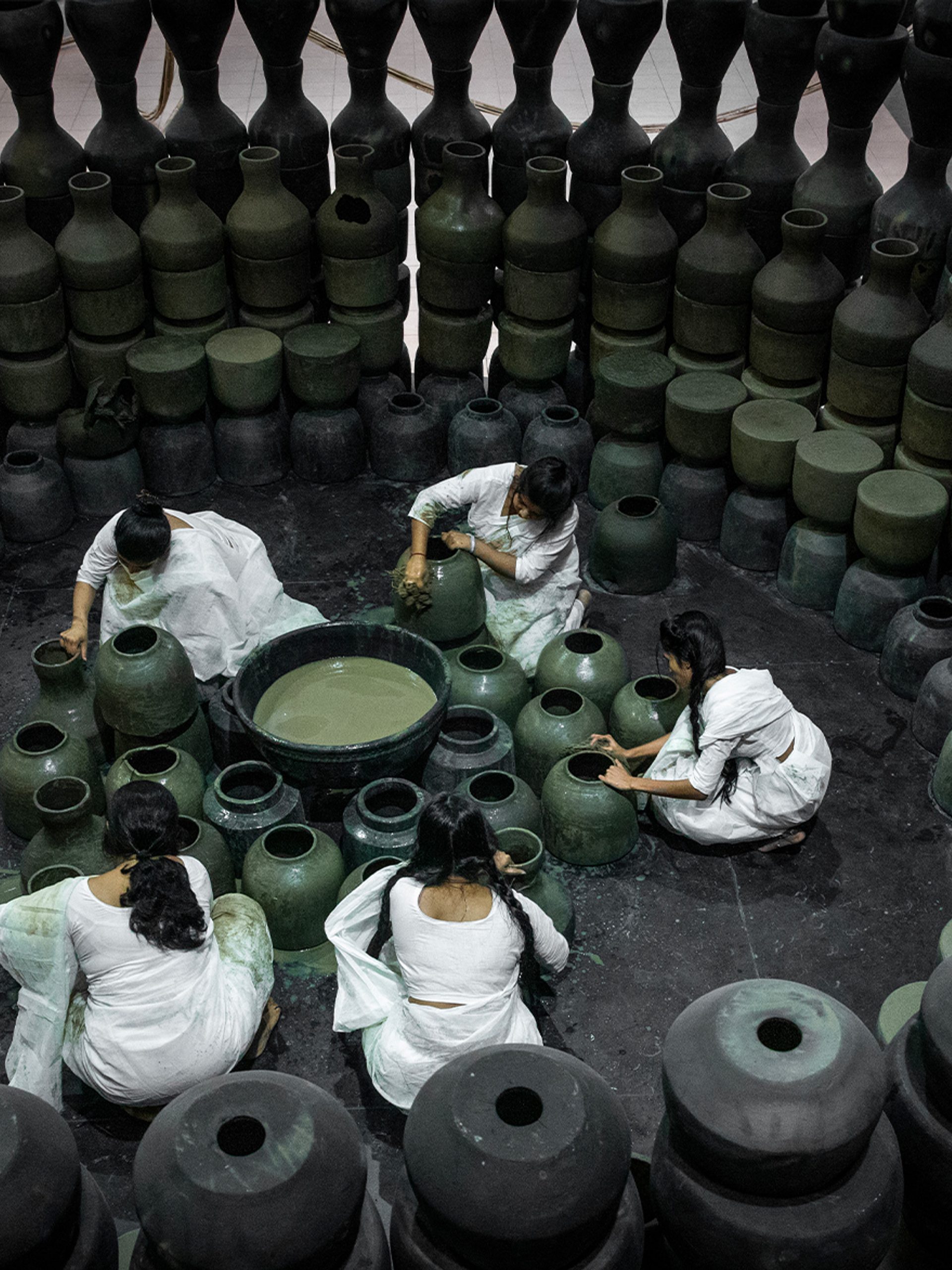 Five people dressed in white sit on the floor around black pots and surrounded by black pots.