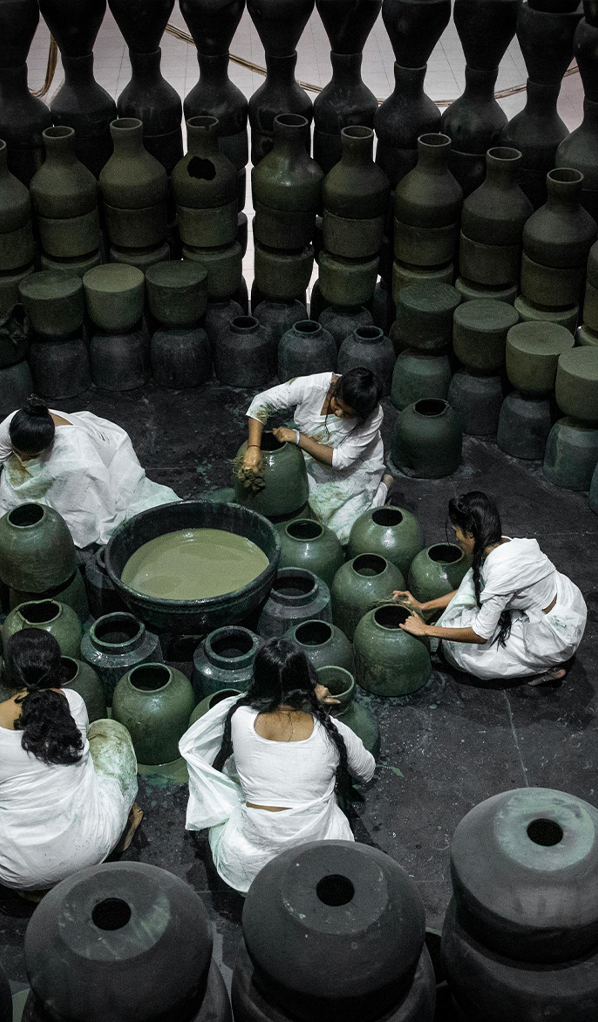 Five people dressed in white sit on the floor around black pots and surrounded by black pots.