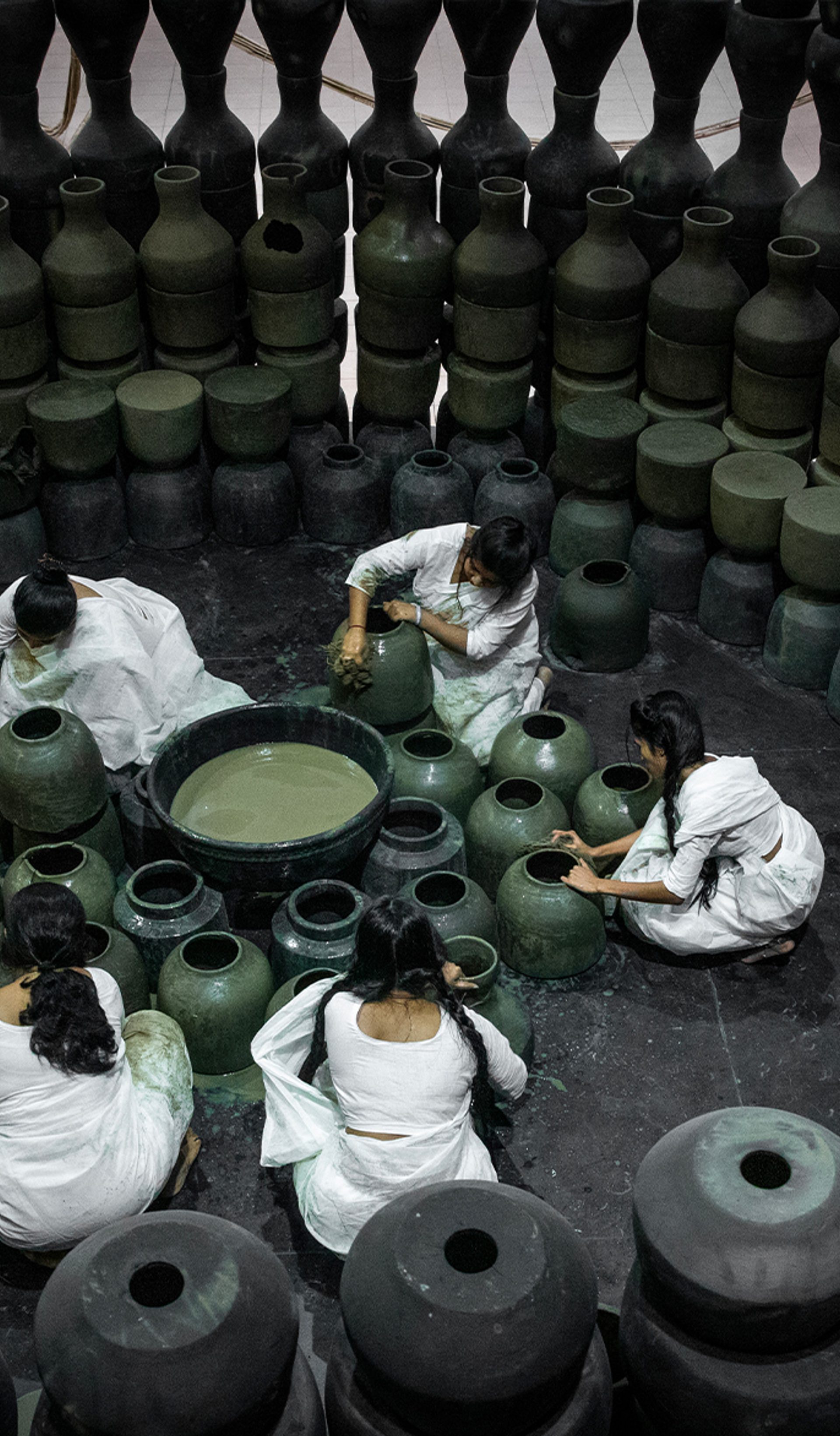Five people dressed in white sit on the floor around black pots and surrounded by black pots.