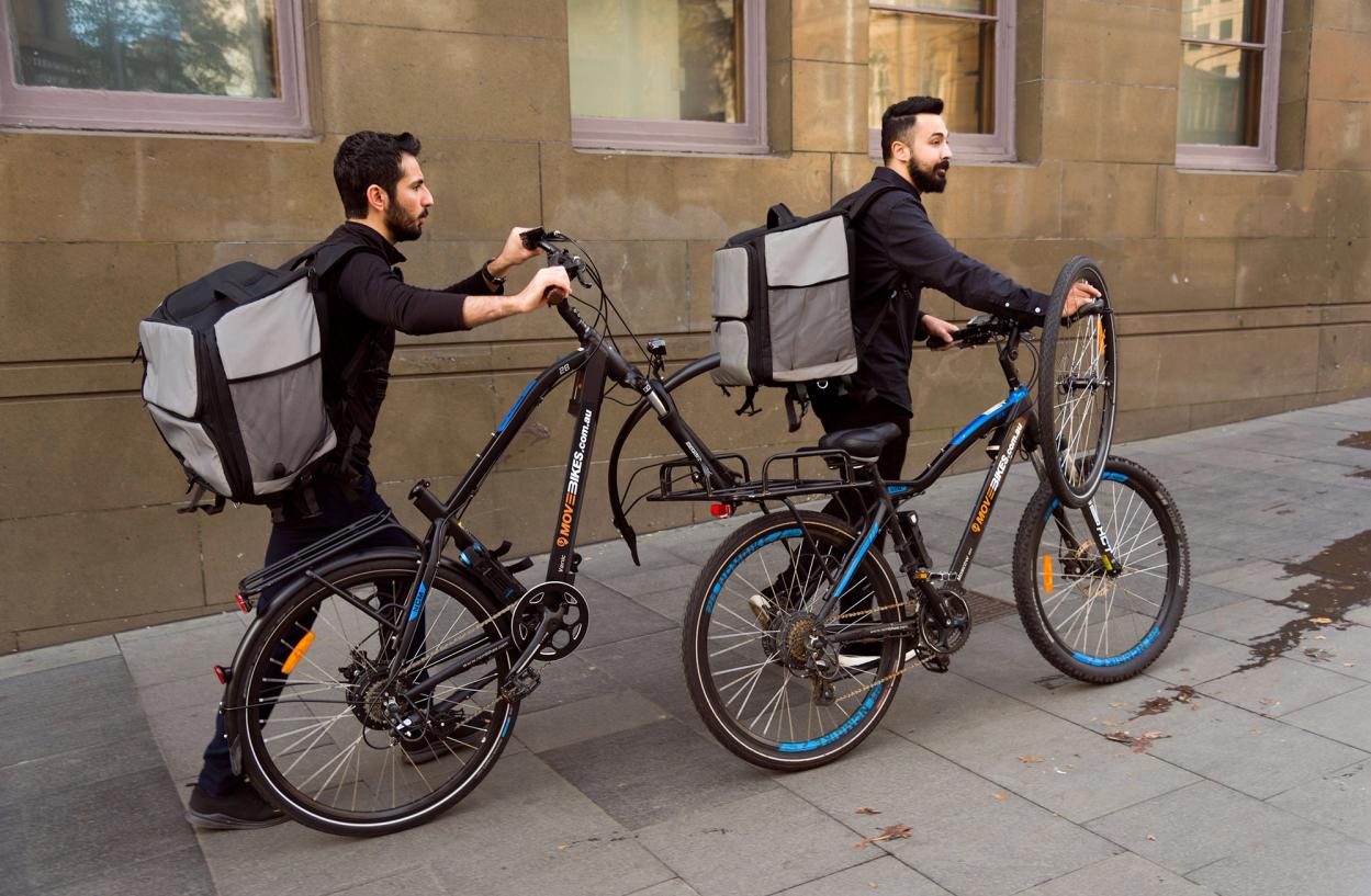 Two figures with food delivery backpacks both dressed in black push their blue bikes along the footpath.