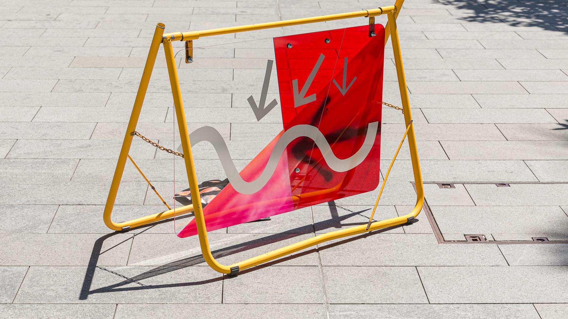 A yellow metal frame on a grey footpath holds up a red and grey sign of arrows and squiggles.