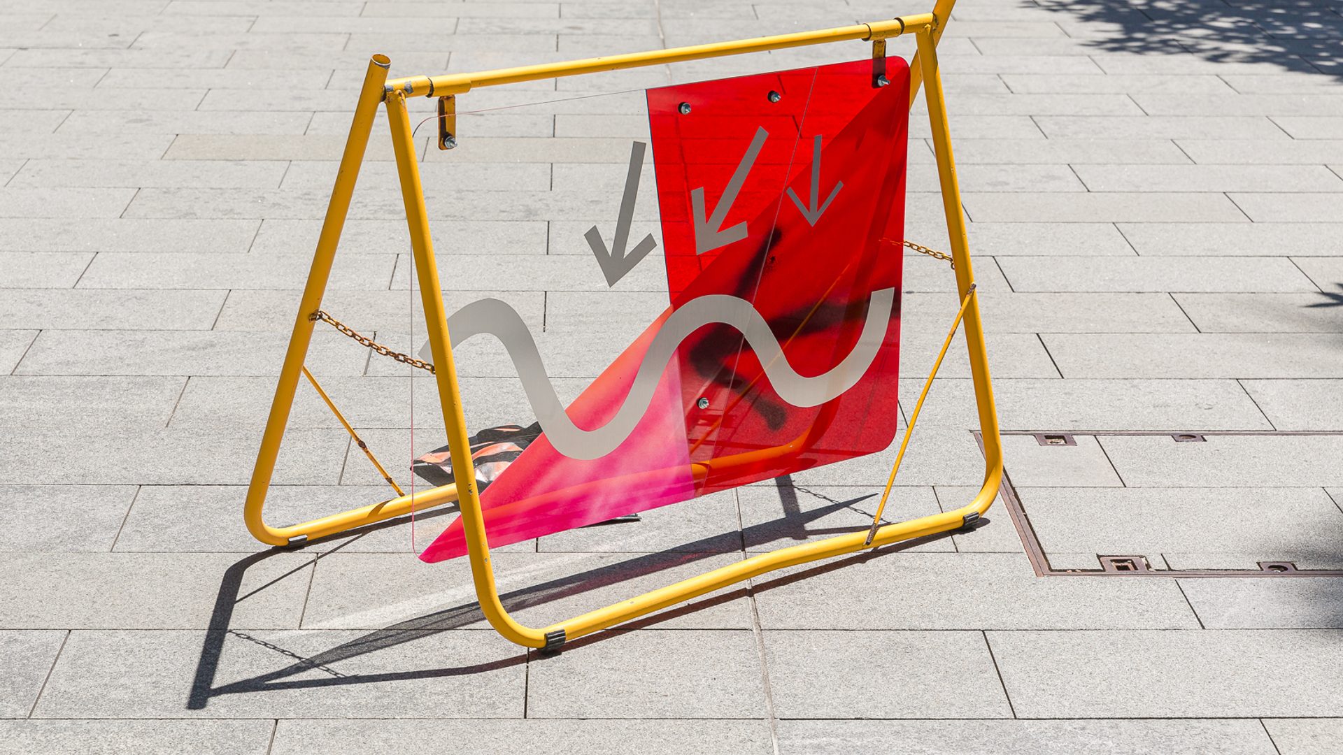A yellow metal frame on a grey footpath holds up a red and grey sign of arrows and squiggles.