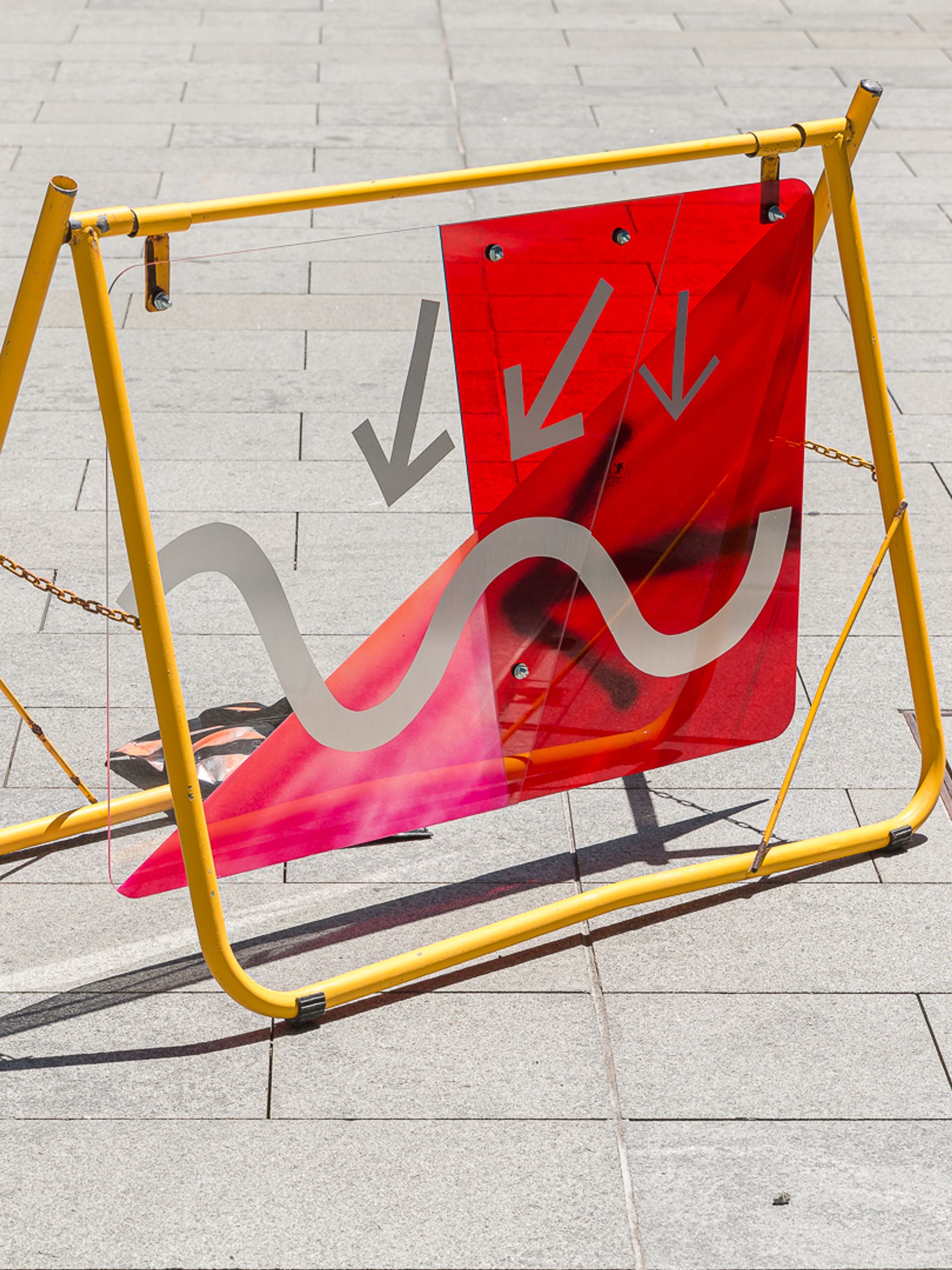 A yellow metal frame on a grey footpath holds up a red and grey sign of arrows and squiggles.