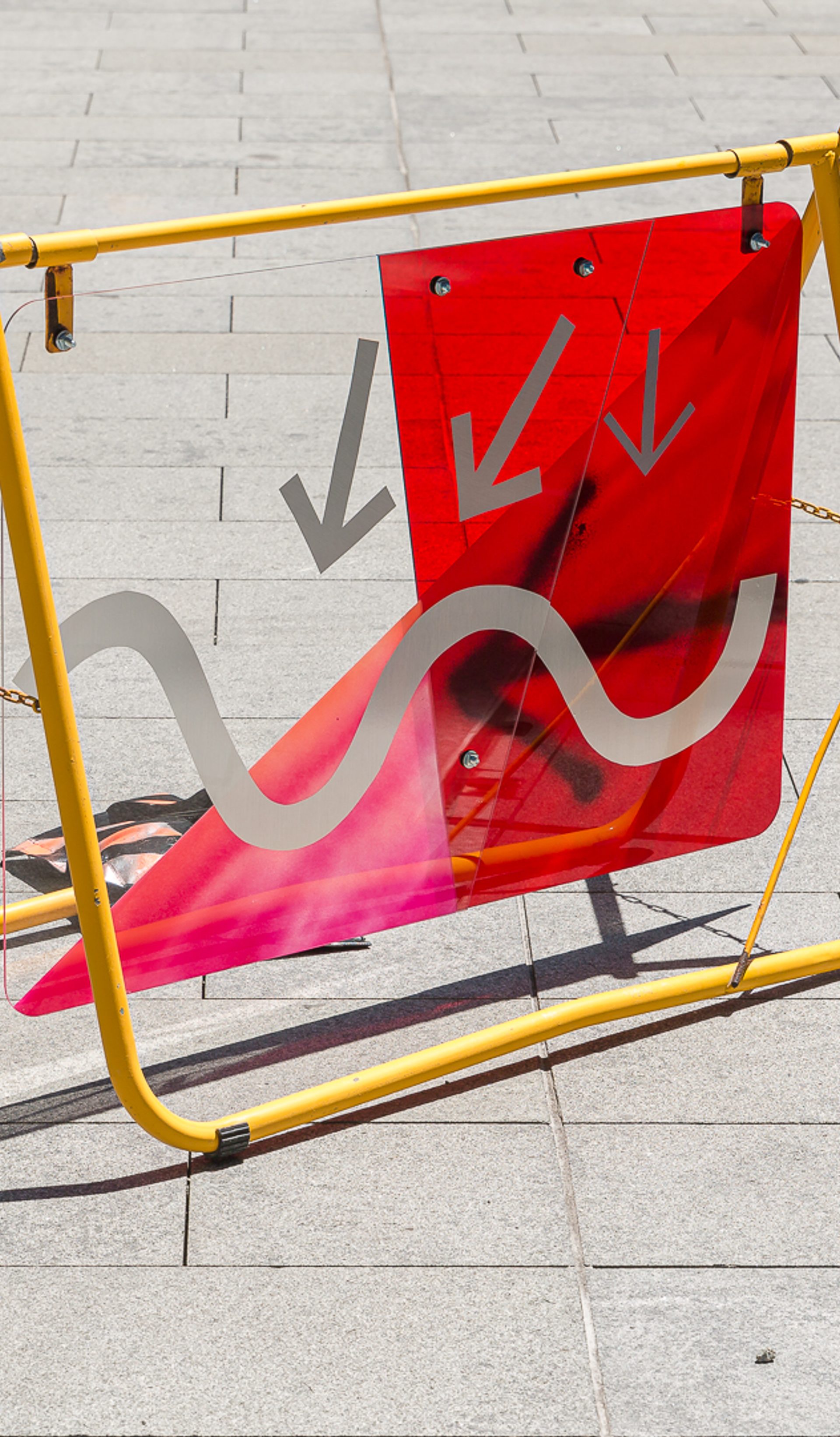 A yellow metal frame on a grey footpath holds up a red and grey sign of arrows and squiggles.