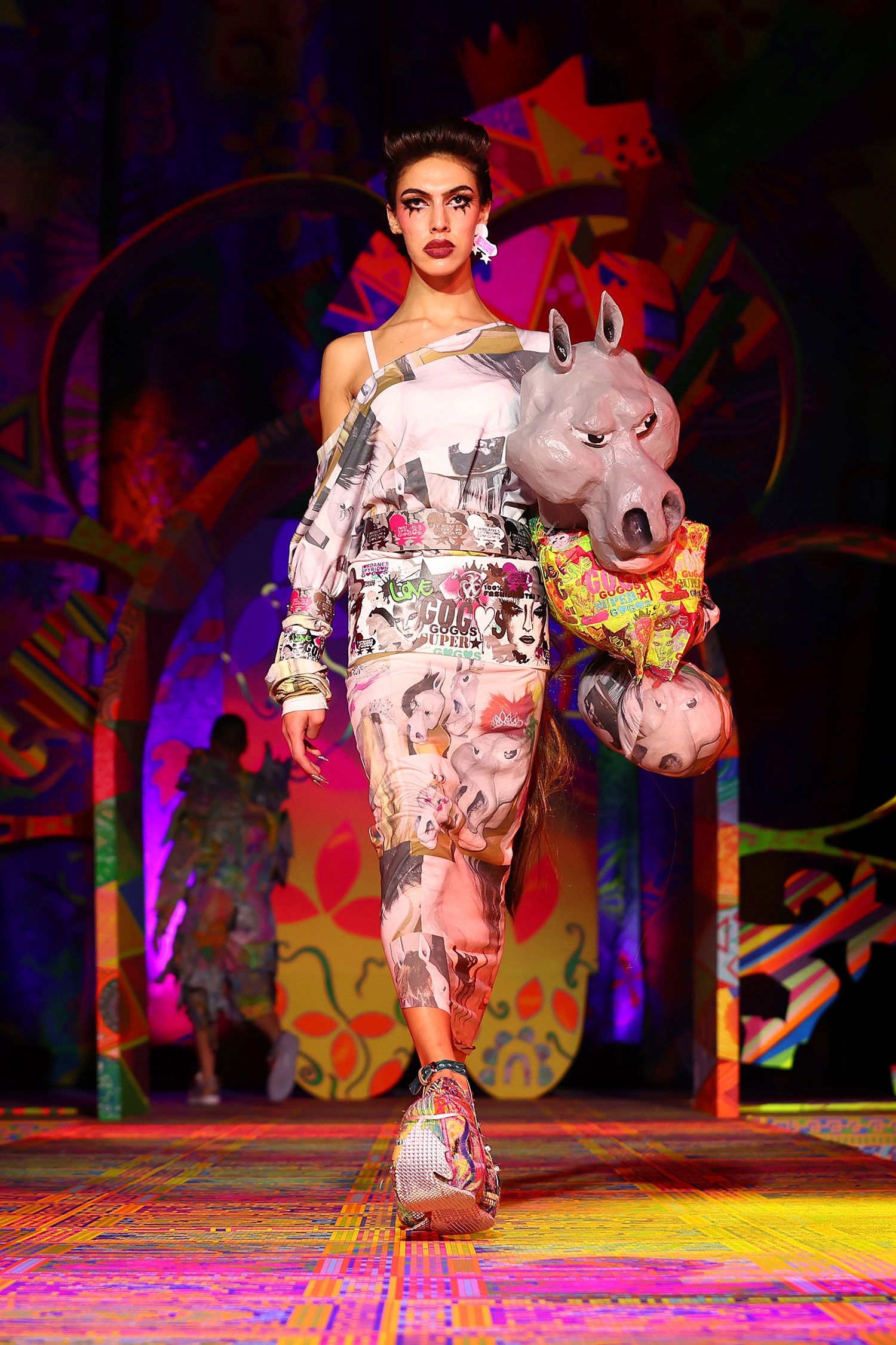 A model walks on the runway with a sculpture of a horses head under their arm.