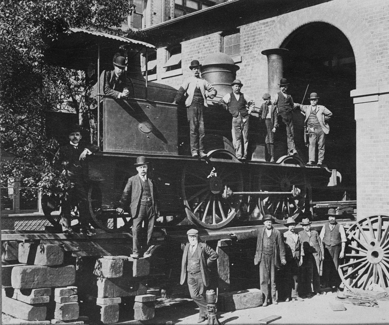 Black and white photograph of men standing on a steam train in front of a brick building